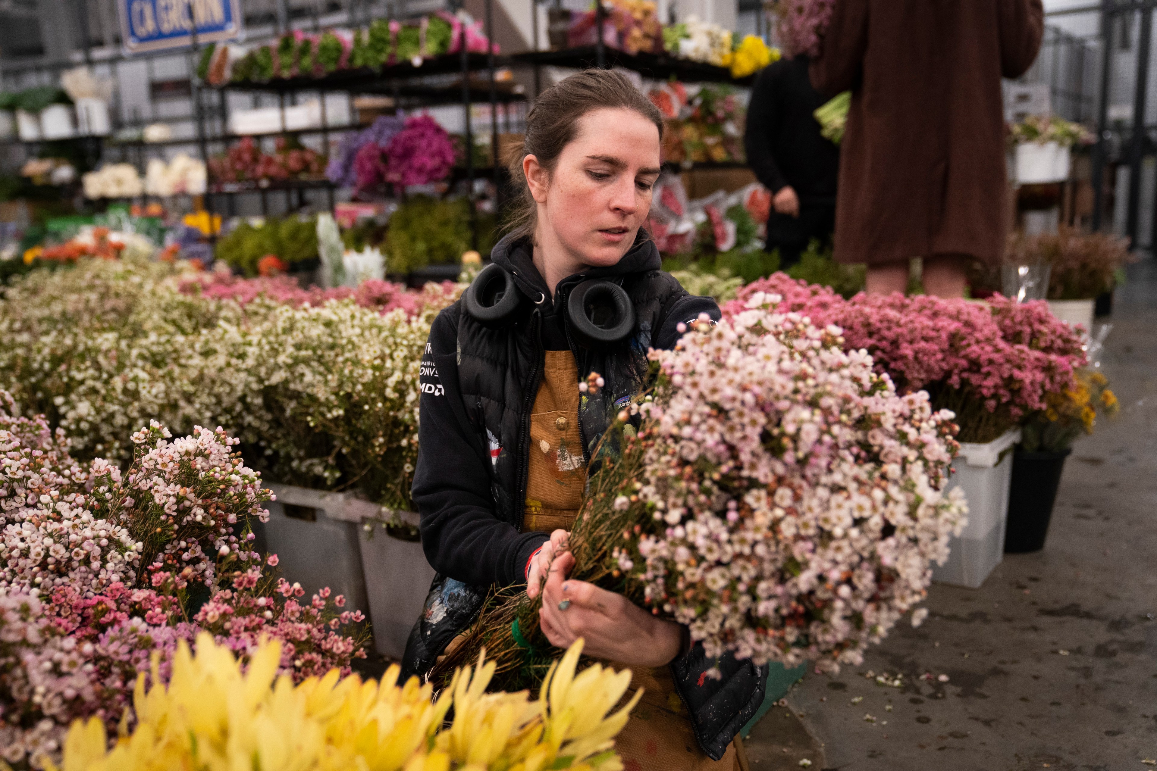 A person in overalls and a black jacket holds a bunch of white and pink flowers in a bustling market, surrounded by vibrant flower arrangements.