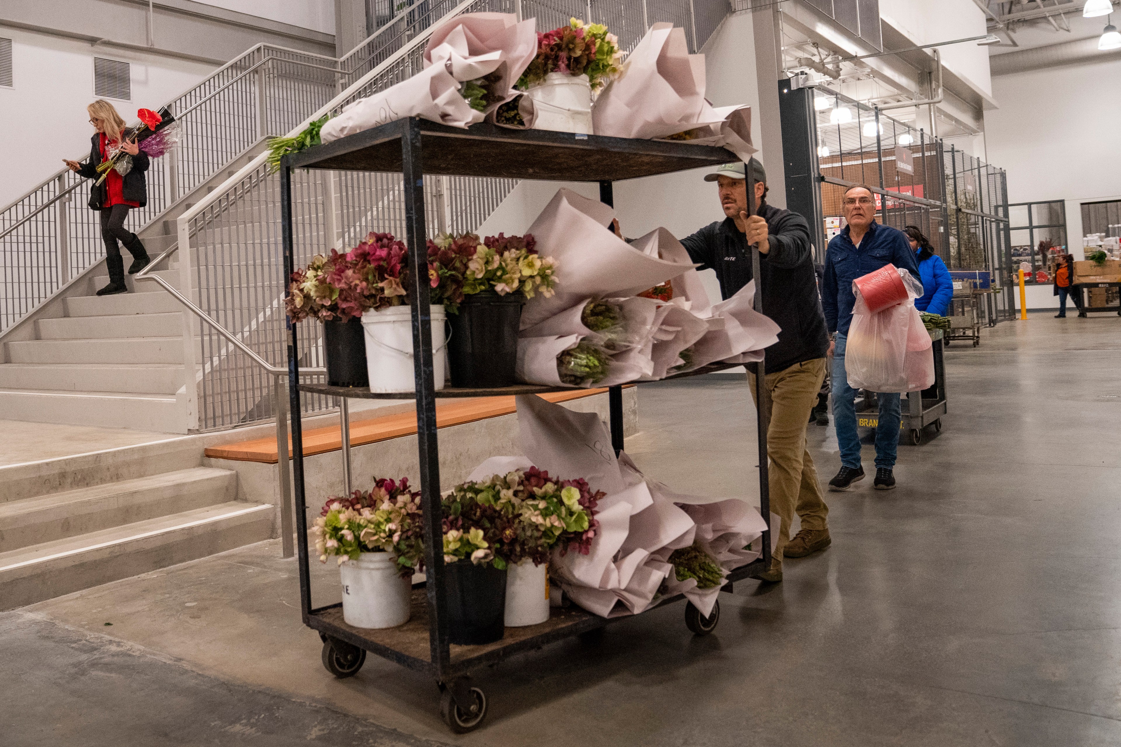 A person pushes a cart filled with buckets of wrapped flowers. Nearby, another person walks down stairs holding flowers and a phone.