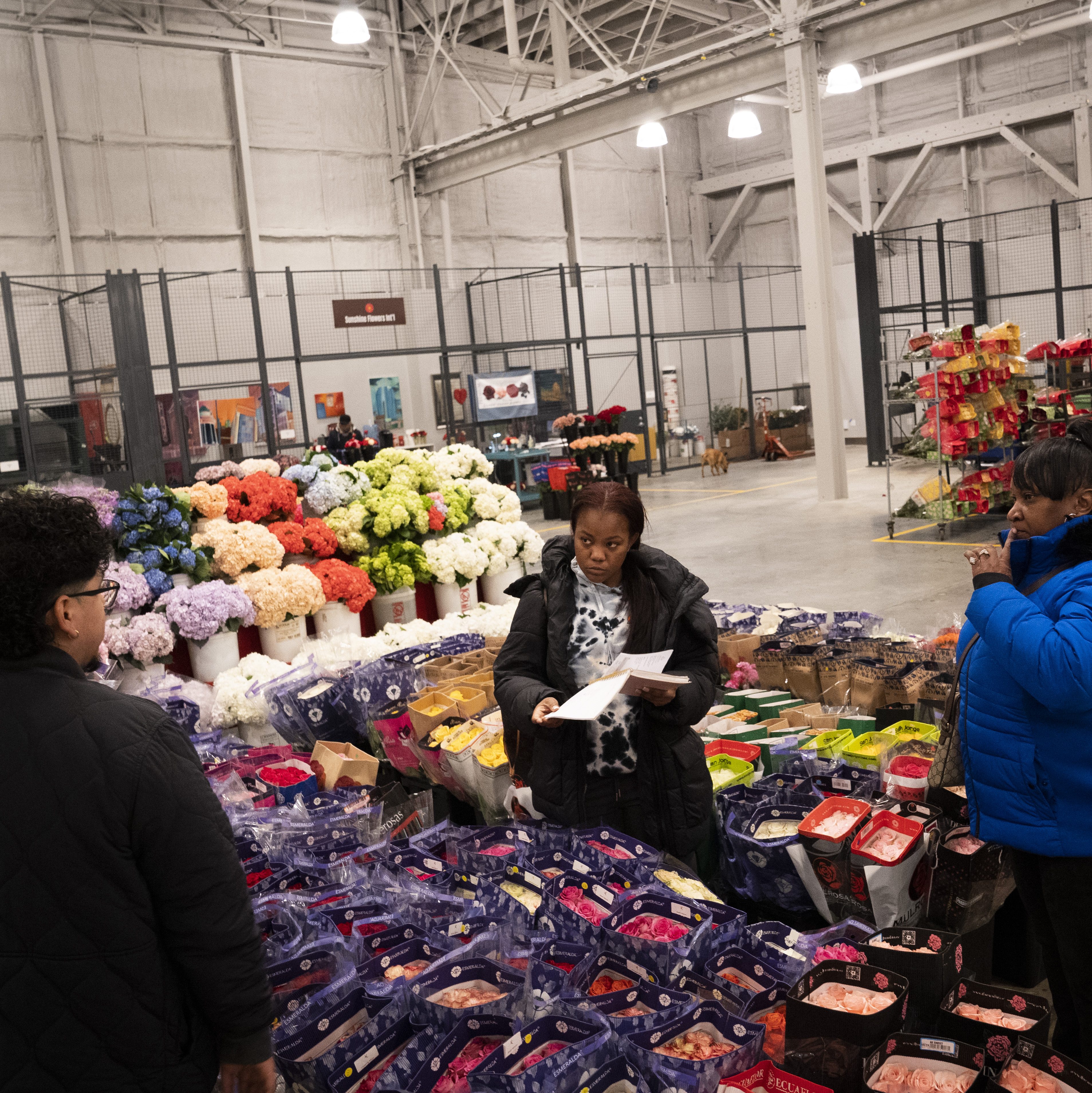 People are standing in a spacious flower market with various colorful bouquets displayed. One person is holding papers, possibly a list or order.