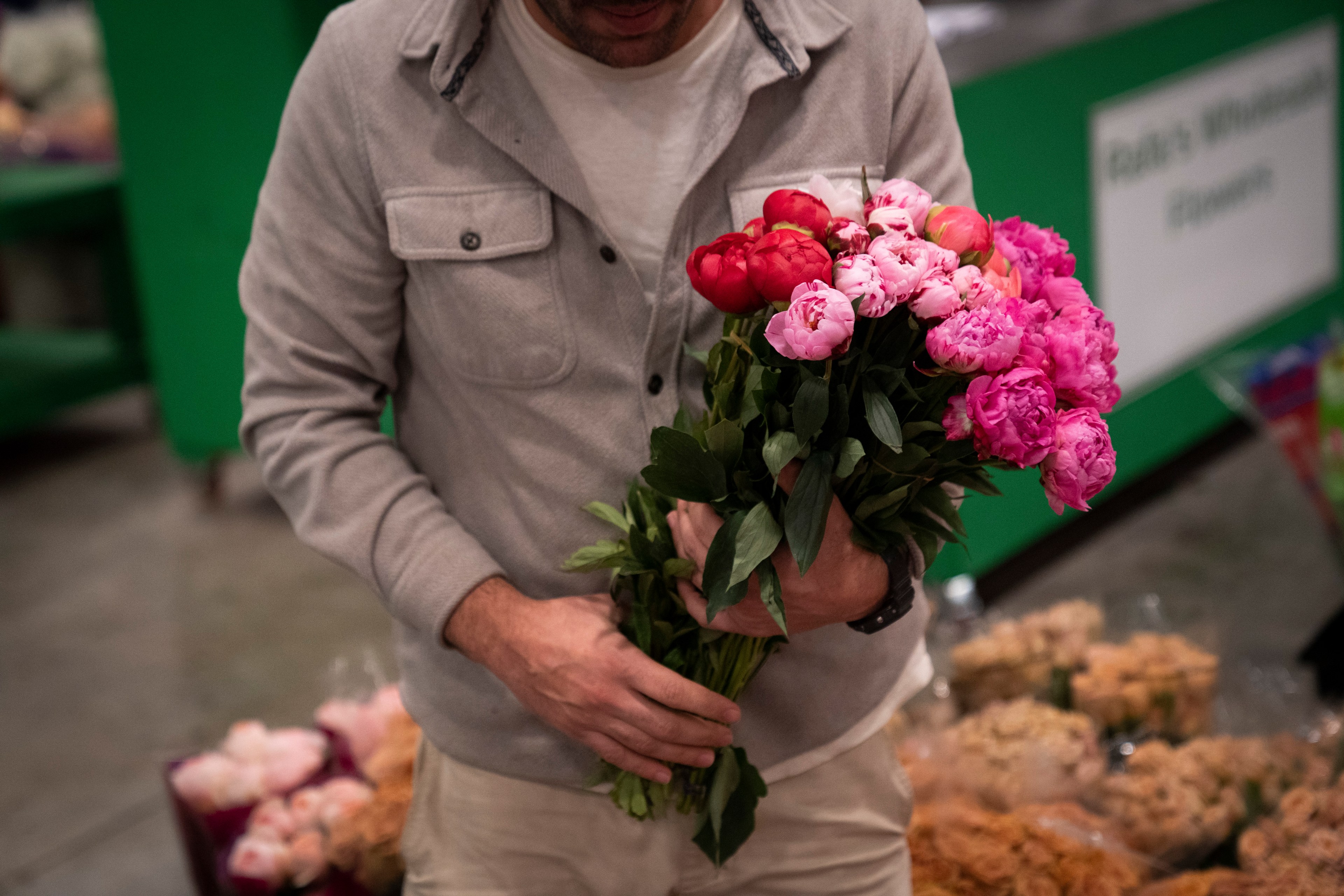 A person in a gray jacket holds a bouquet of pink and red peonies, standing in a market setting with colorful flower bouquets in the background.