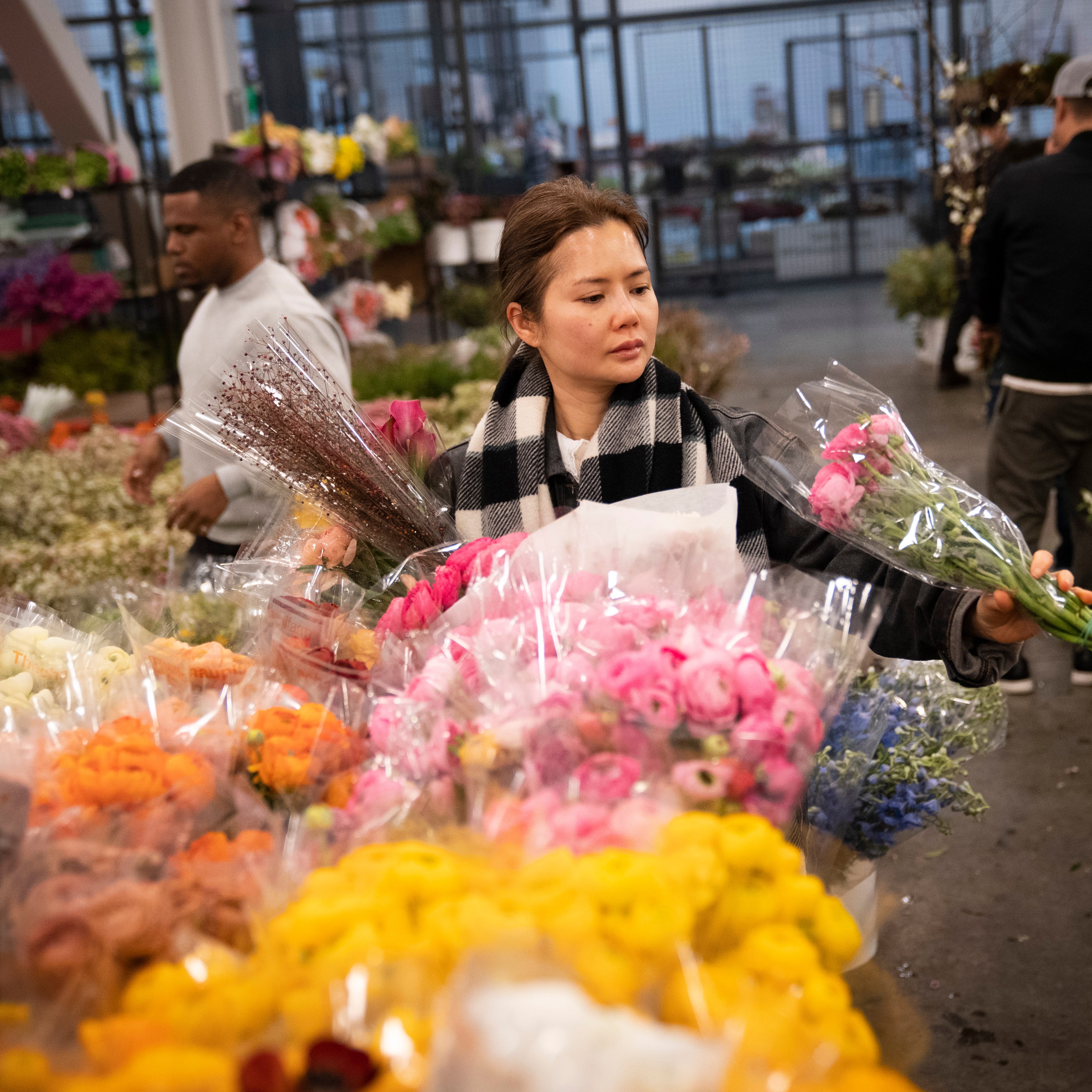 A woman in a scarf selects pink flowers in a bustling flower market, surrounded by colorful bouquets and bustling shoppers in the background.
