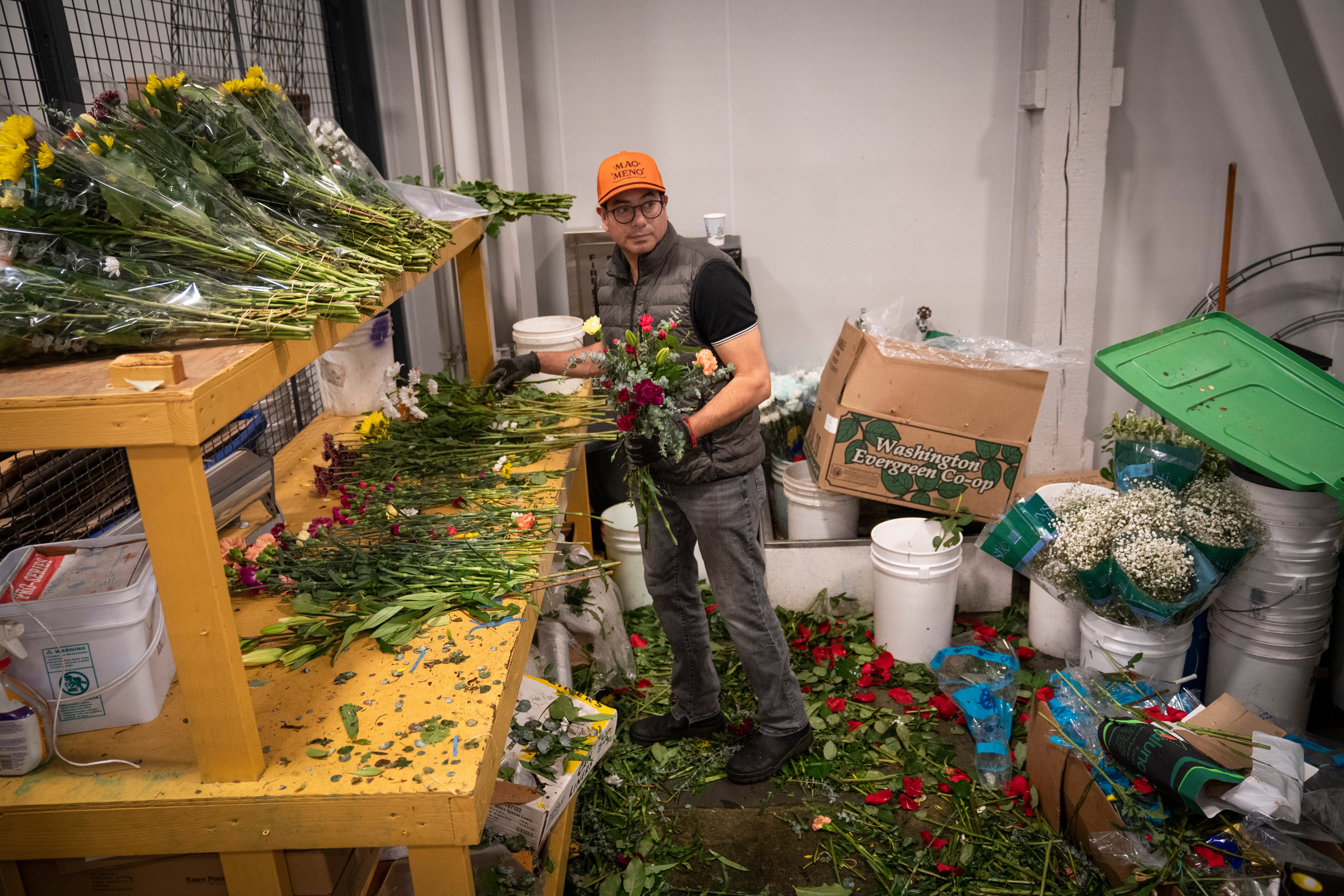 A person stands in a workshop arranging flowers, surrounded by shelves of floral bundles and scattered petals. They wear an orange cap and a dark vest.