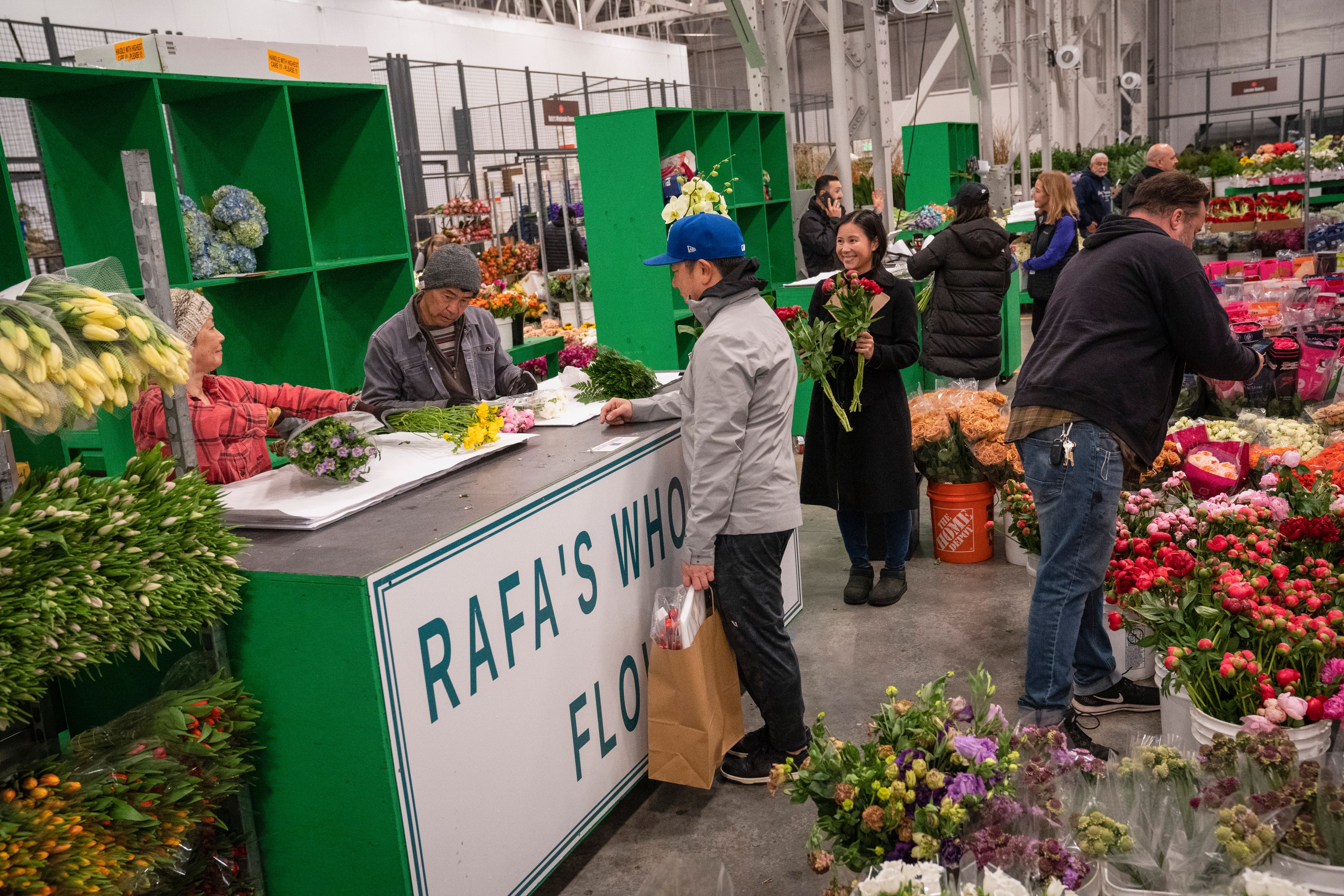 People are buying flowers at a bustling flower market, with vibrant bouquets on display and interactions at a counter labeled &quot;Rafa's Wholesale Flowers.&quot;