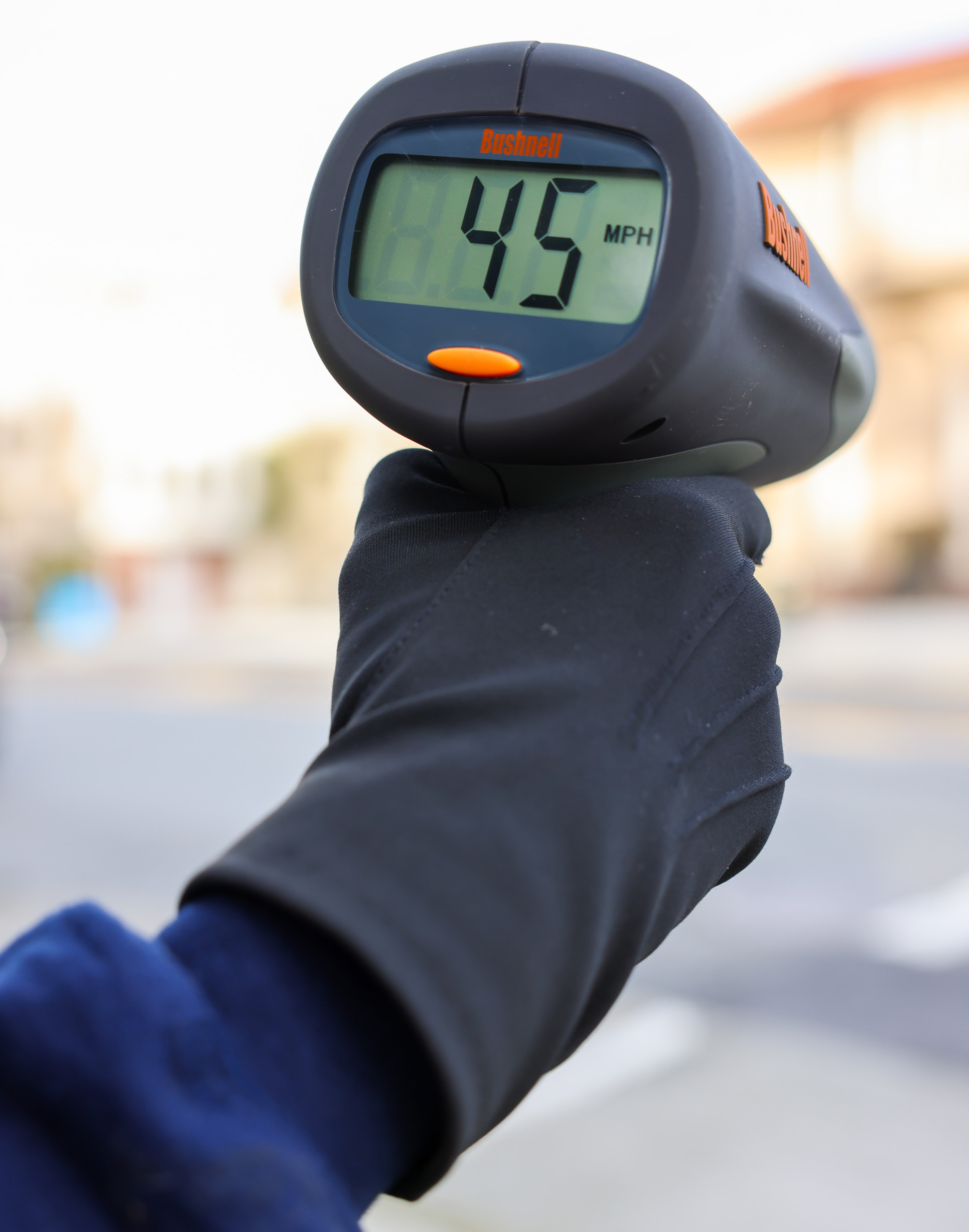 A person wearing a black glove is holding a speed radar gun displaying &quot;45 MPH&quot; against a blurred background.