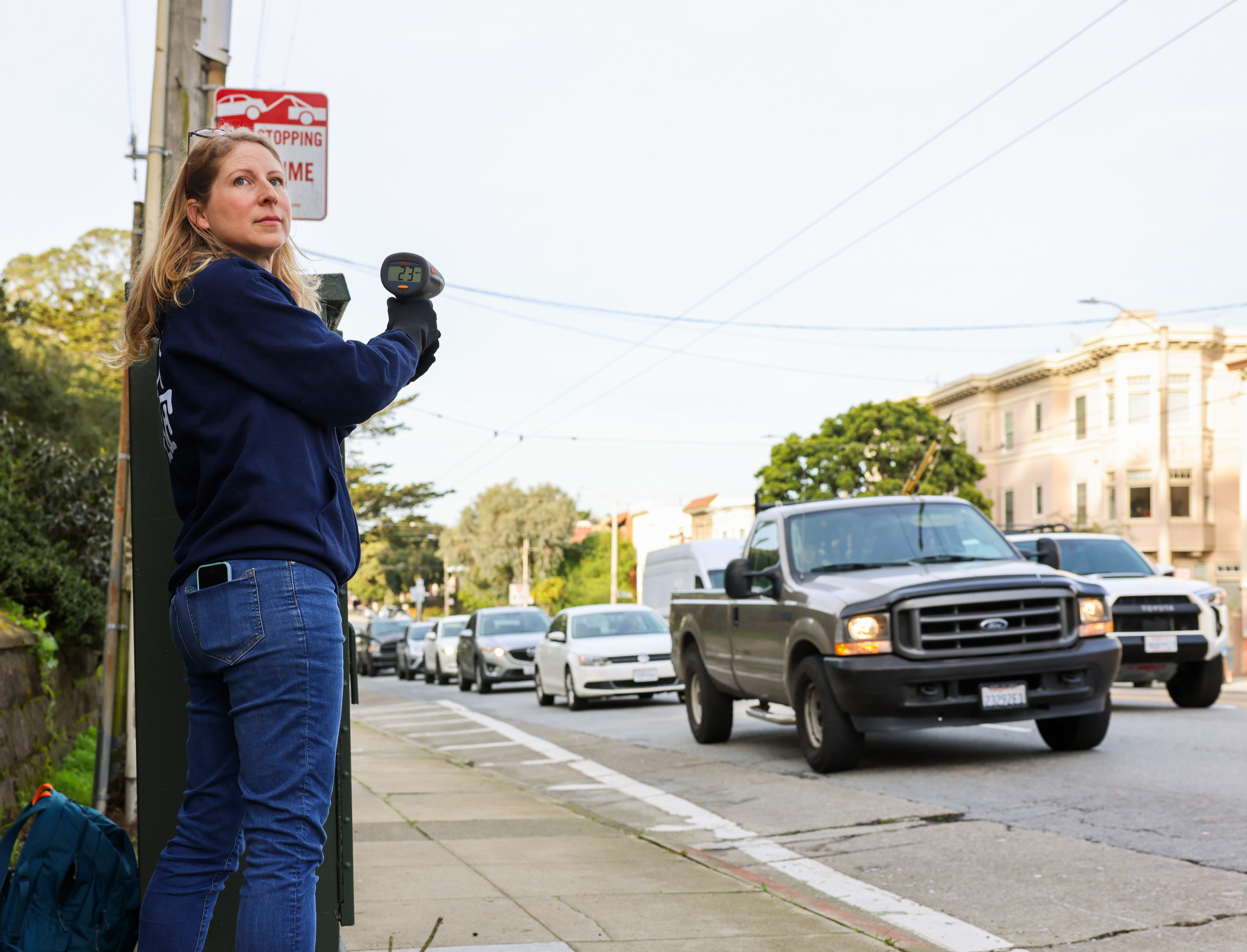 A woman stands on a sidewalk pointing a radar gun at passing vehicles on a busy street. She wears jeans and a blue sweatshirt, with a no stopping sign nearby.