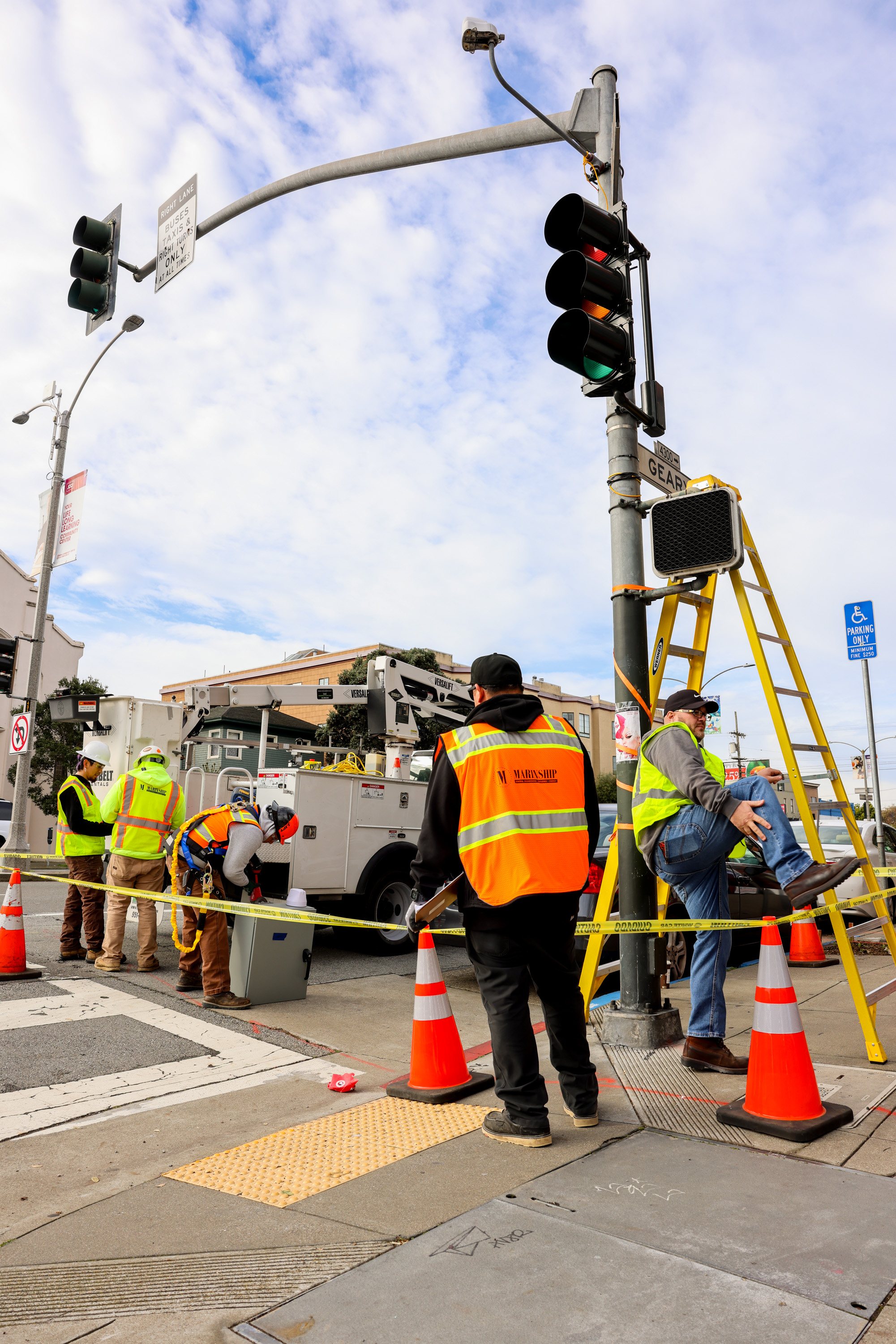 Construction workers in reflective vests are repairing a traffic light at an intersection. Orange cones and caution tape surround the area under a cloudy sky.