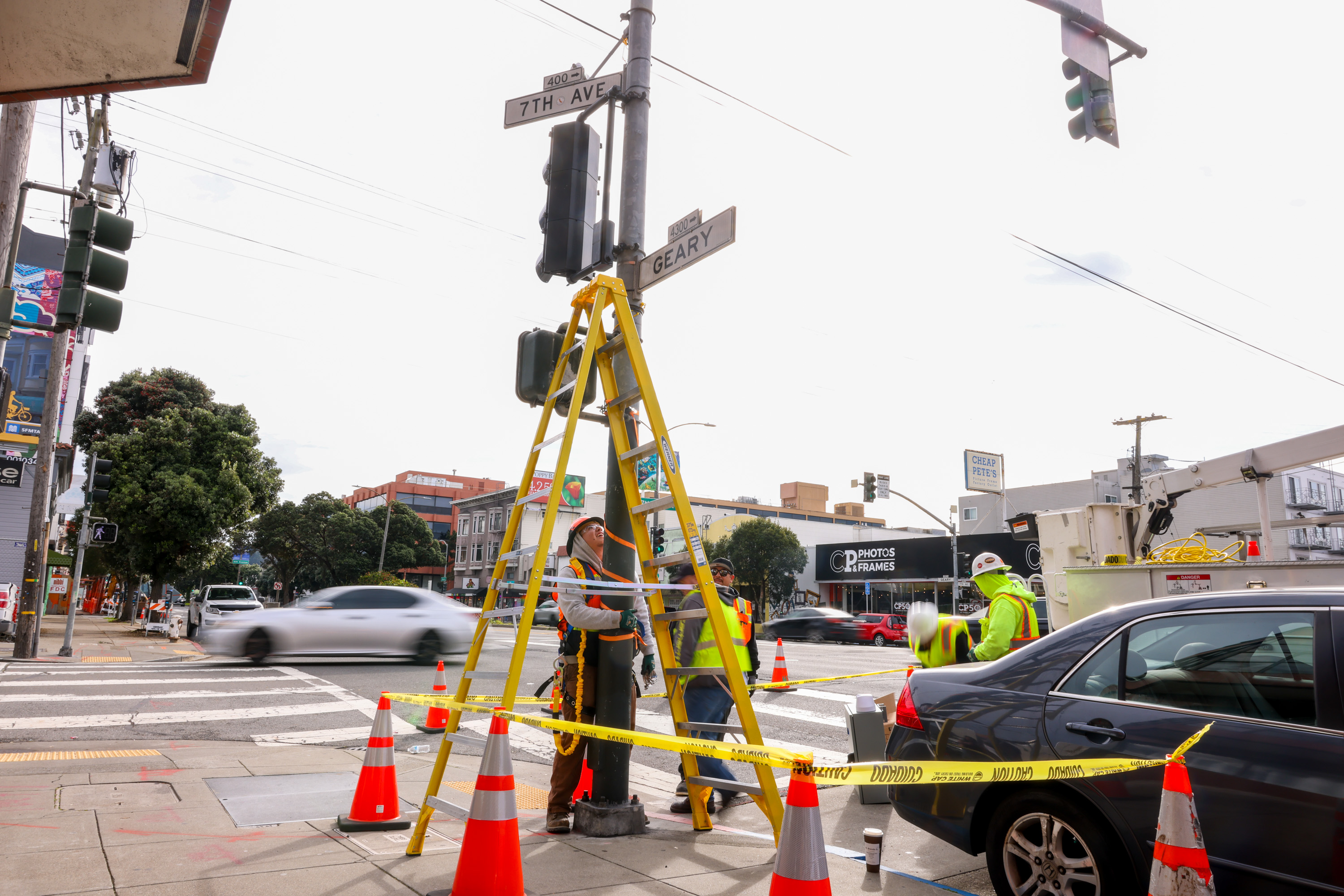 Construction workers in safety gear are standing at a traffic intersection with a ladder propped against a pole, surrounded by cones and caution tape.