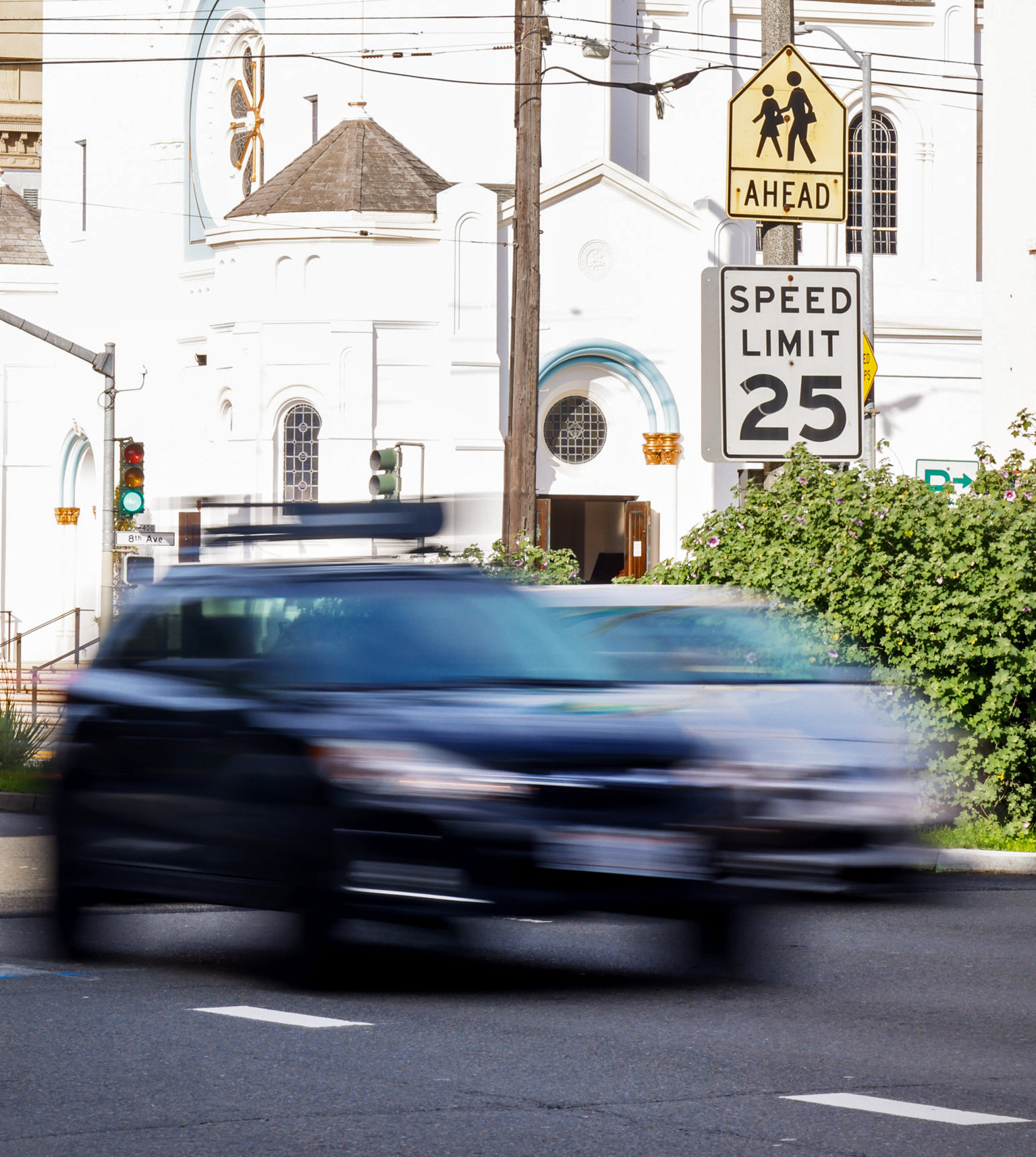 A speeding black car blurs past a white building. A school zone sign and a speed limit sign of 25 mph are visible, with greenery and a traffic light nearby.