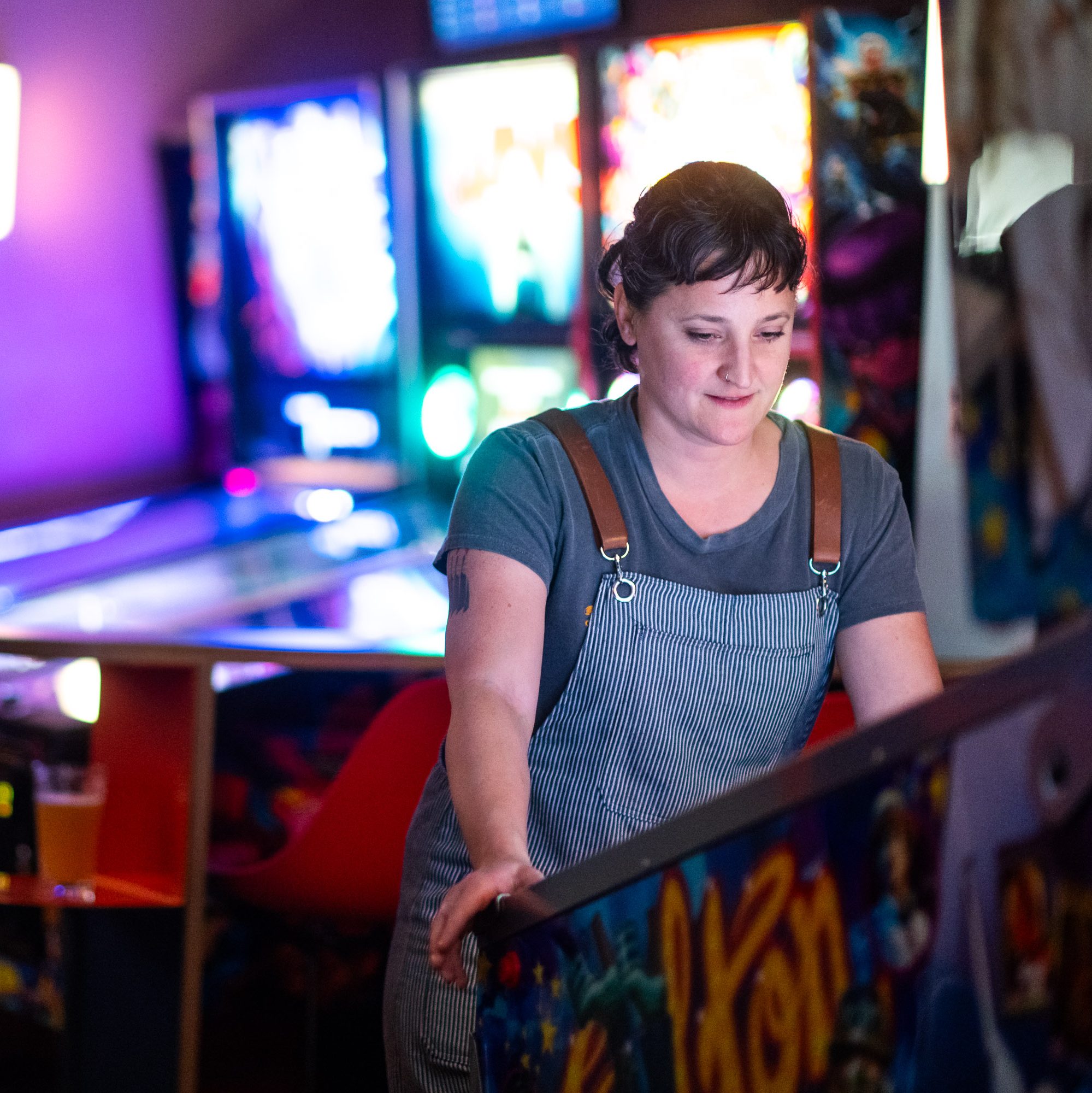 A person is focused on playing a pinball machine in an arcade setting. They're wearing a striped apron over a gray t-shirt. The background is colorful and lit.