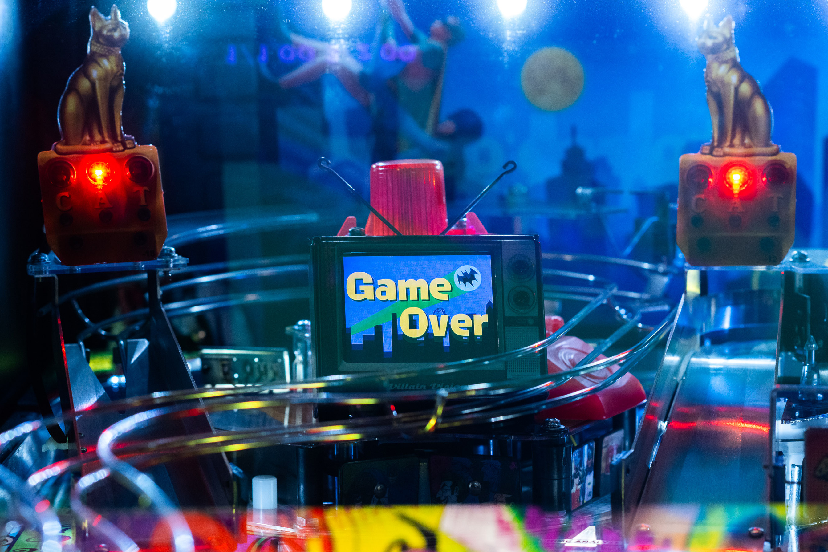 A pinball machine displays "Game Over" on a screen. Two cat figures with glowing red eyes sit atop light fixtures. The background features a cityscape and moon.