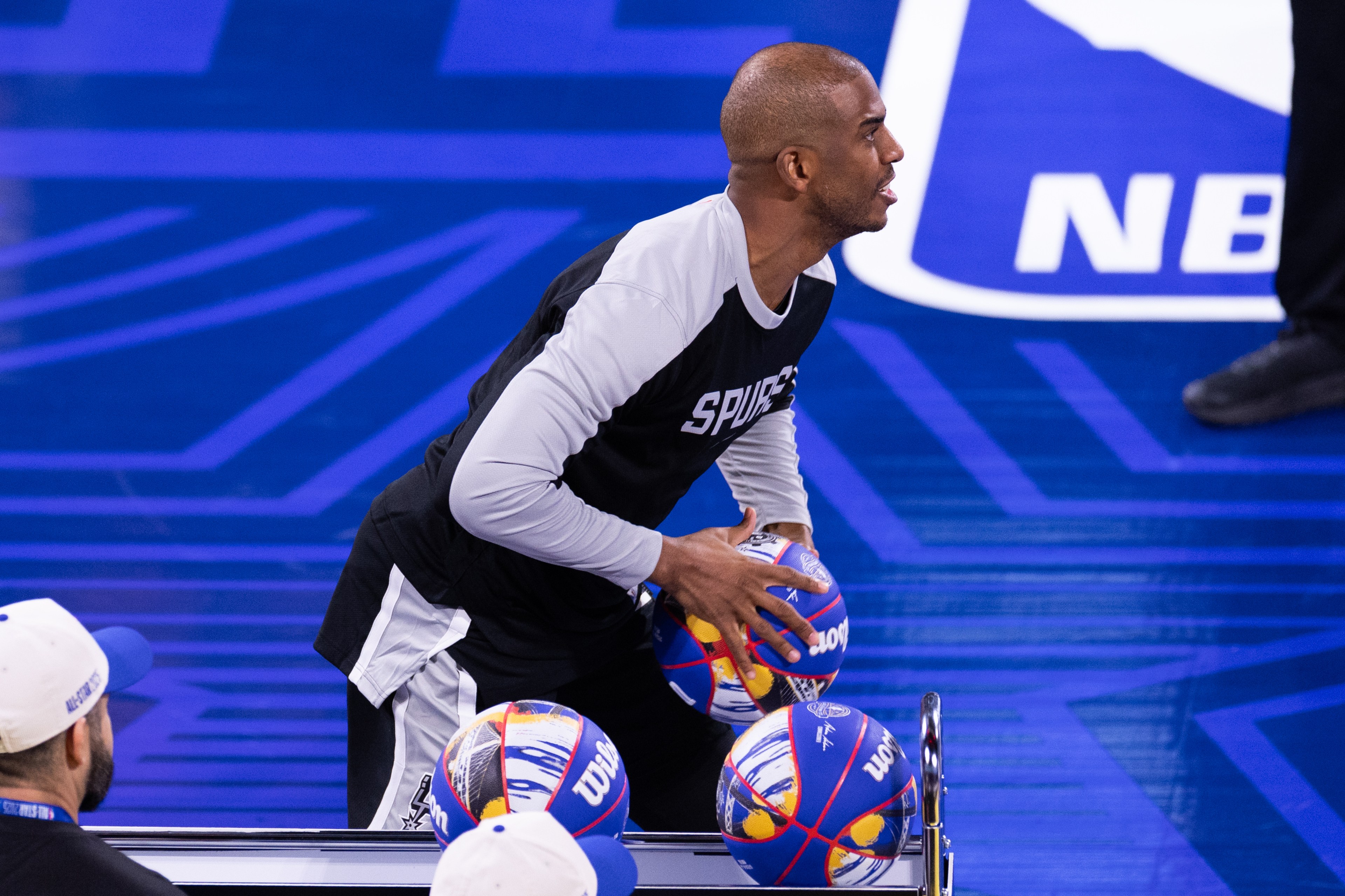 A person in a &quot;Spurs&quot; shirt is holding colorful basketballs on a court with an NBA logo, preparing for an activity.