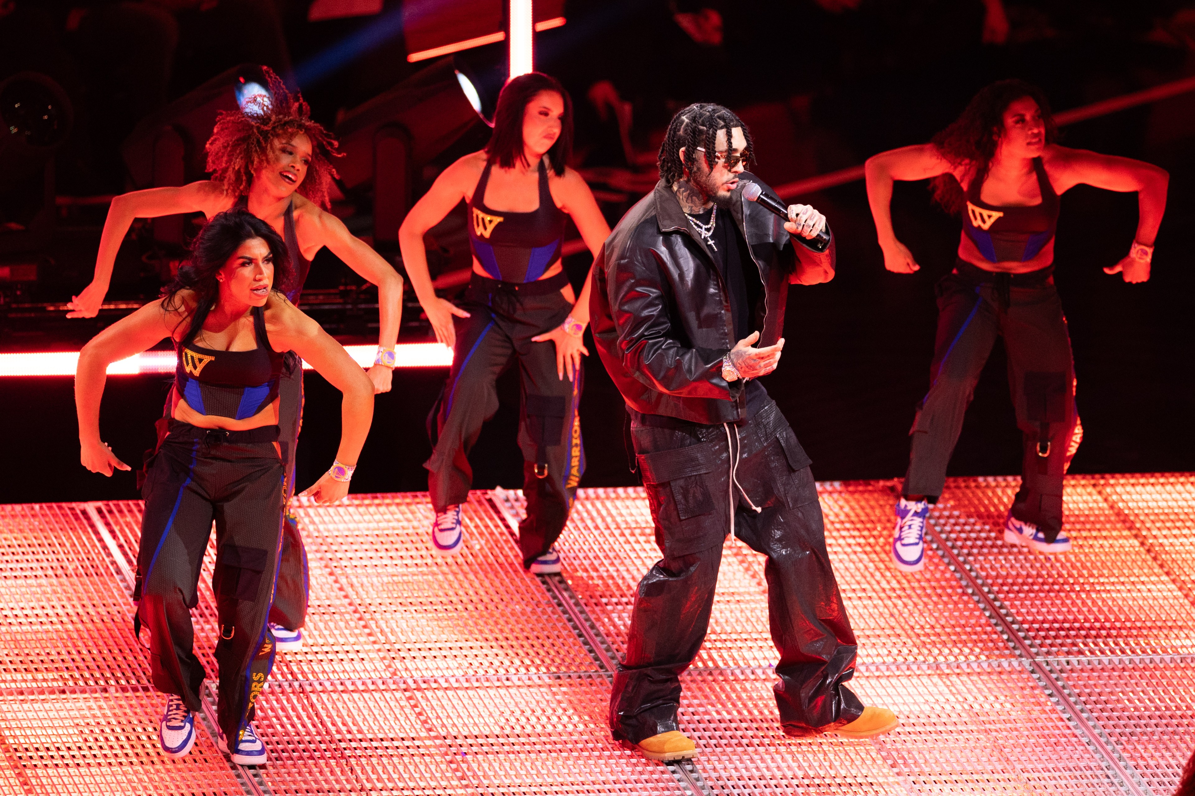 A performer in a black leather outfit sings into a microphone on a lit stage. Four dancers in matching athletic wear dance energetically behind him.