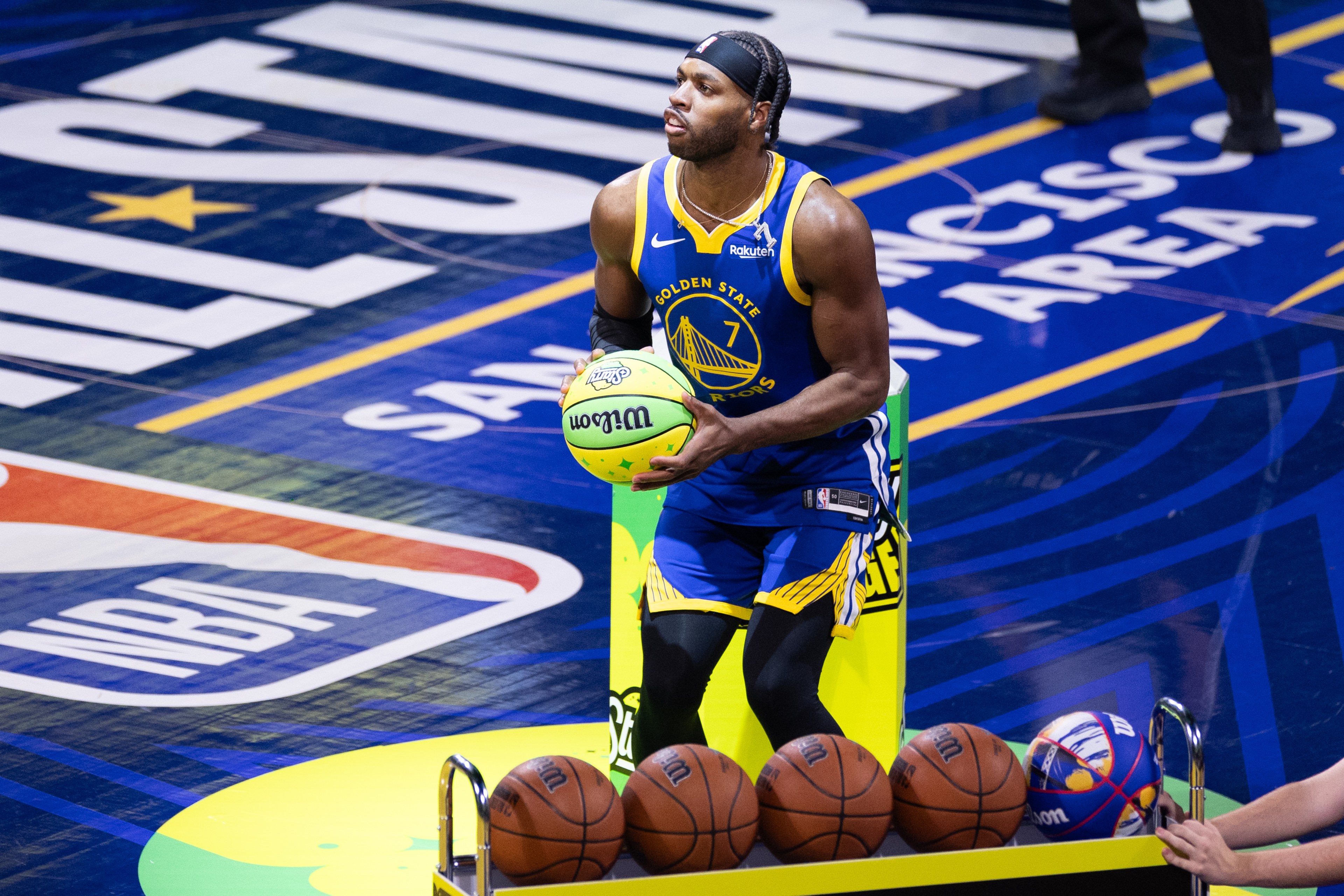 A basketball player in a Golden State Warriors jersey holds a neon green basketball, preparing to shoot. Several other basketballs are on a rack beside him.