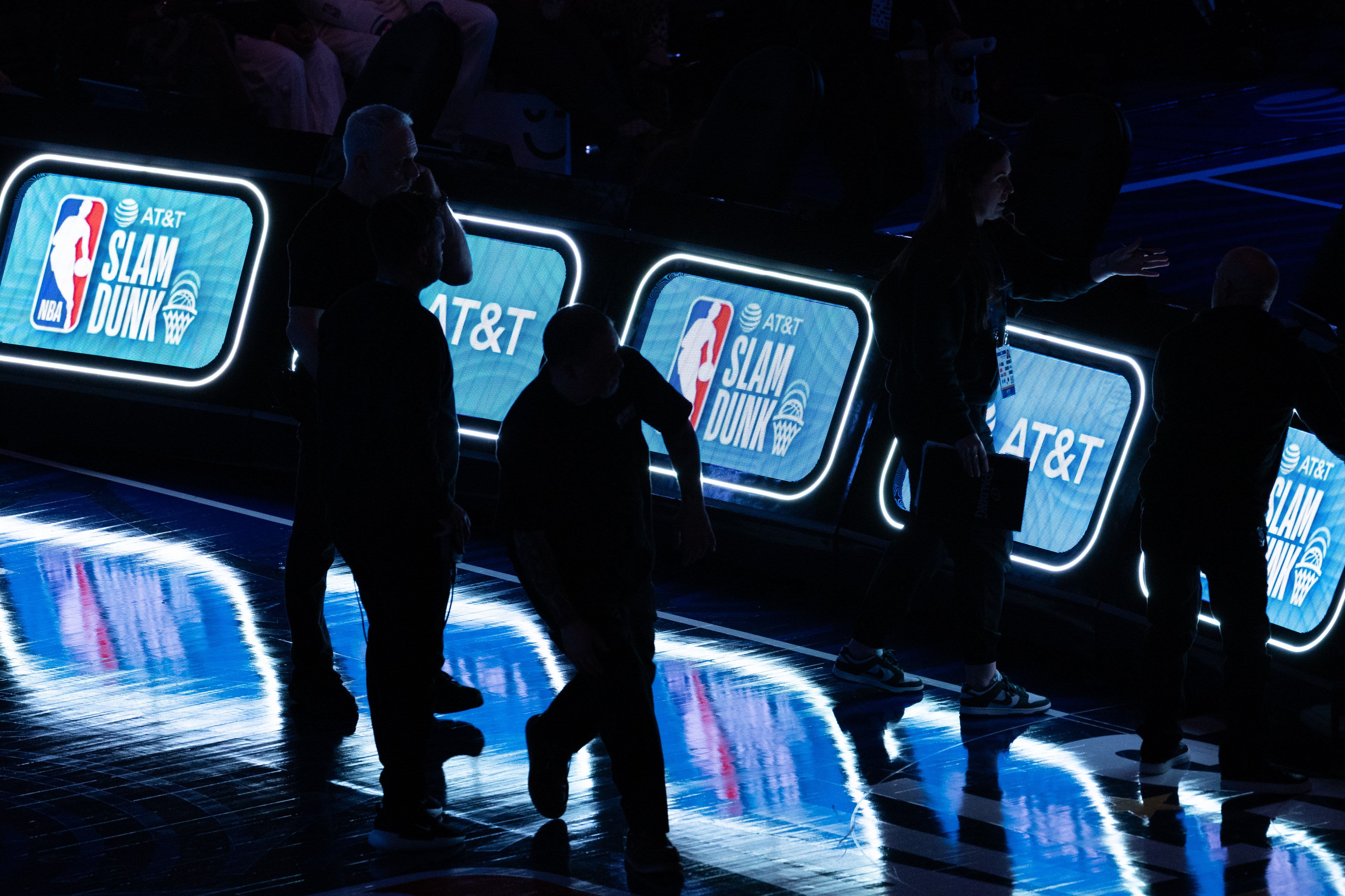 Several people stand in a dimly lit area with illuminated panels displaying &quot;NBA AT&amp;T Slam Dunk,&quot; casting blue reflections on the shiny floor.