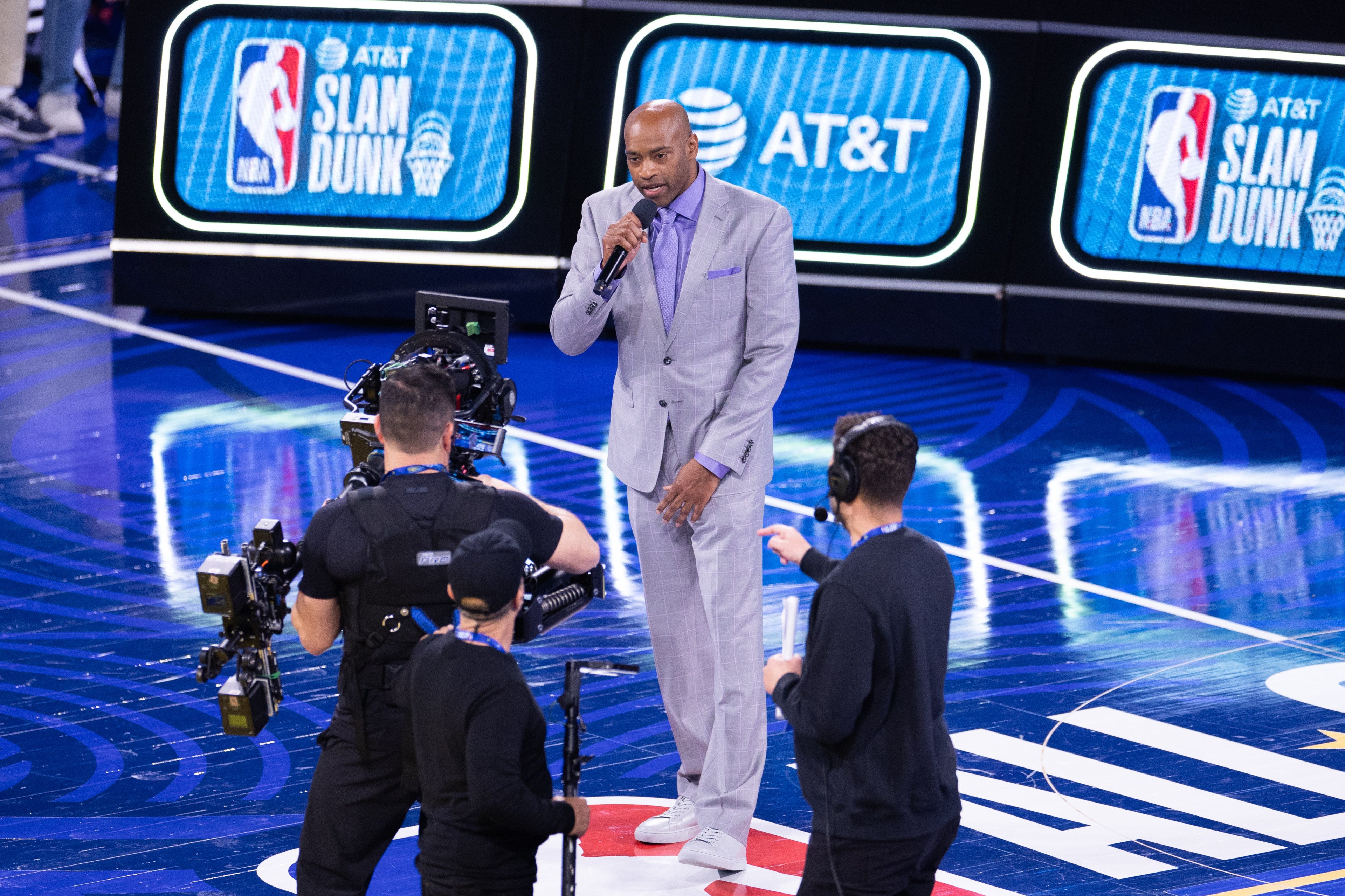 A man in a light gray suit is speaking into a microphone on a basketball court. There are cameras and crew around him, with &quot;NBA Slam Dunk&quot; signs in the background.