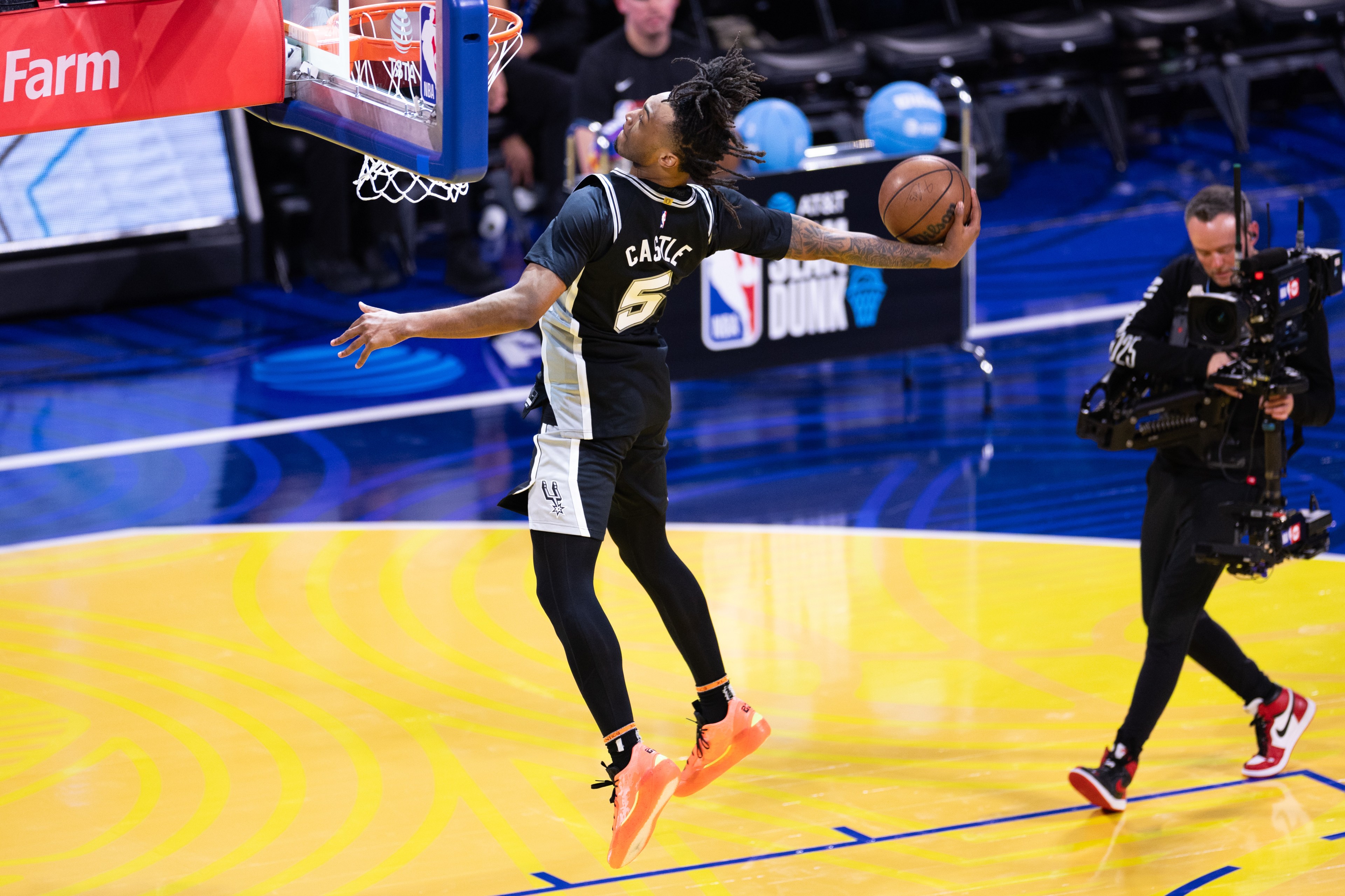 A basketball player is mid-air, about to perform a dunk, with one hand holding the ball. A cameraman films the action on the court.