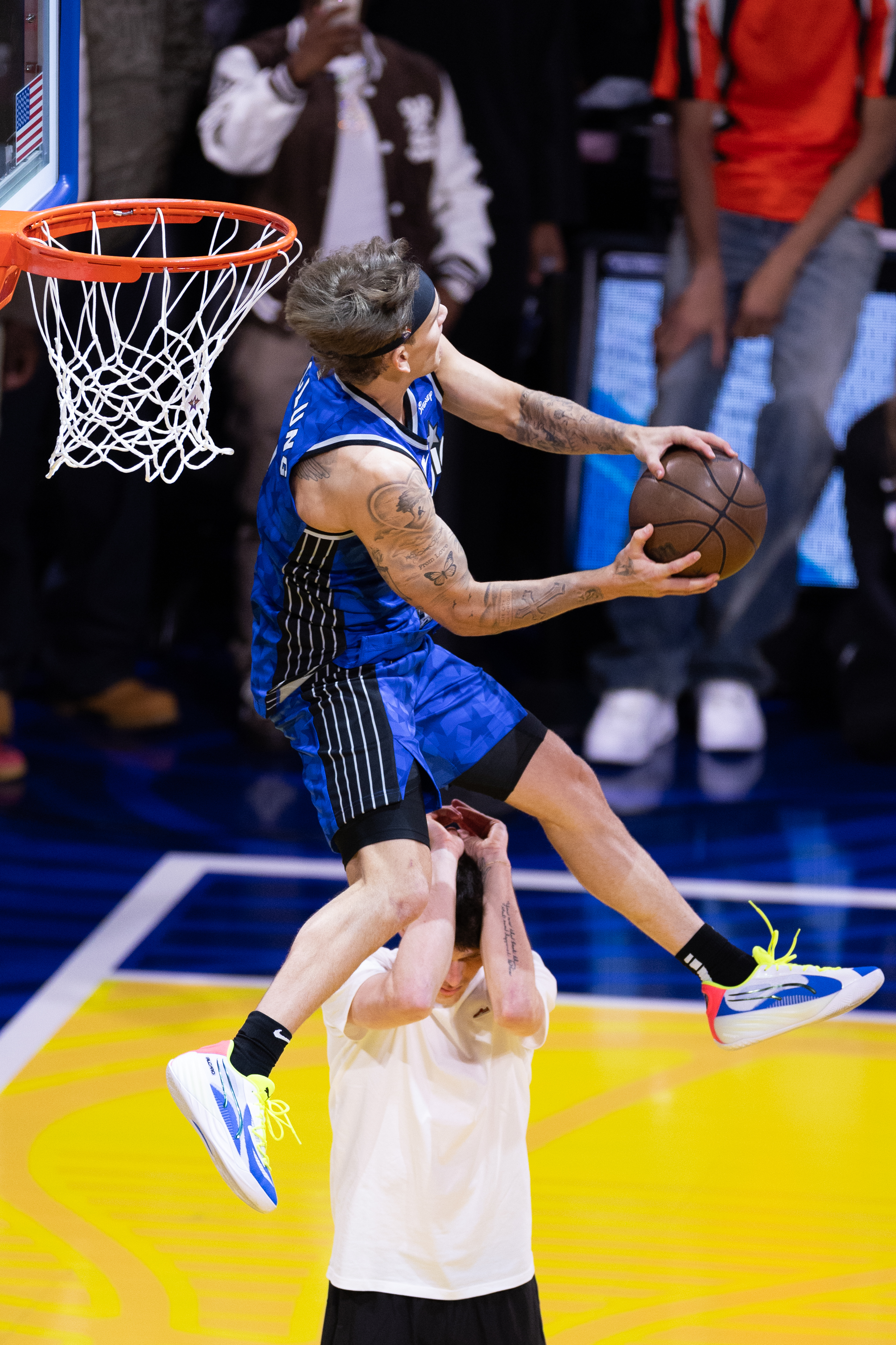 A basketball player in a blue jersey is performing a jump, holding a ball near the hoop, with another person standing beneath him.