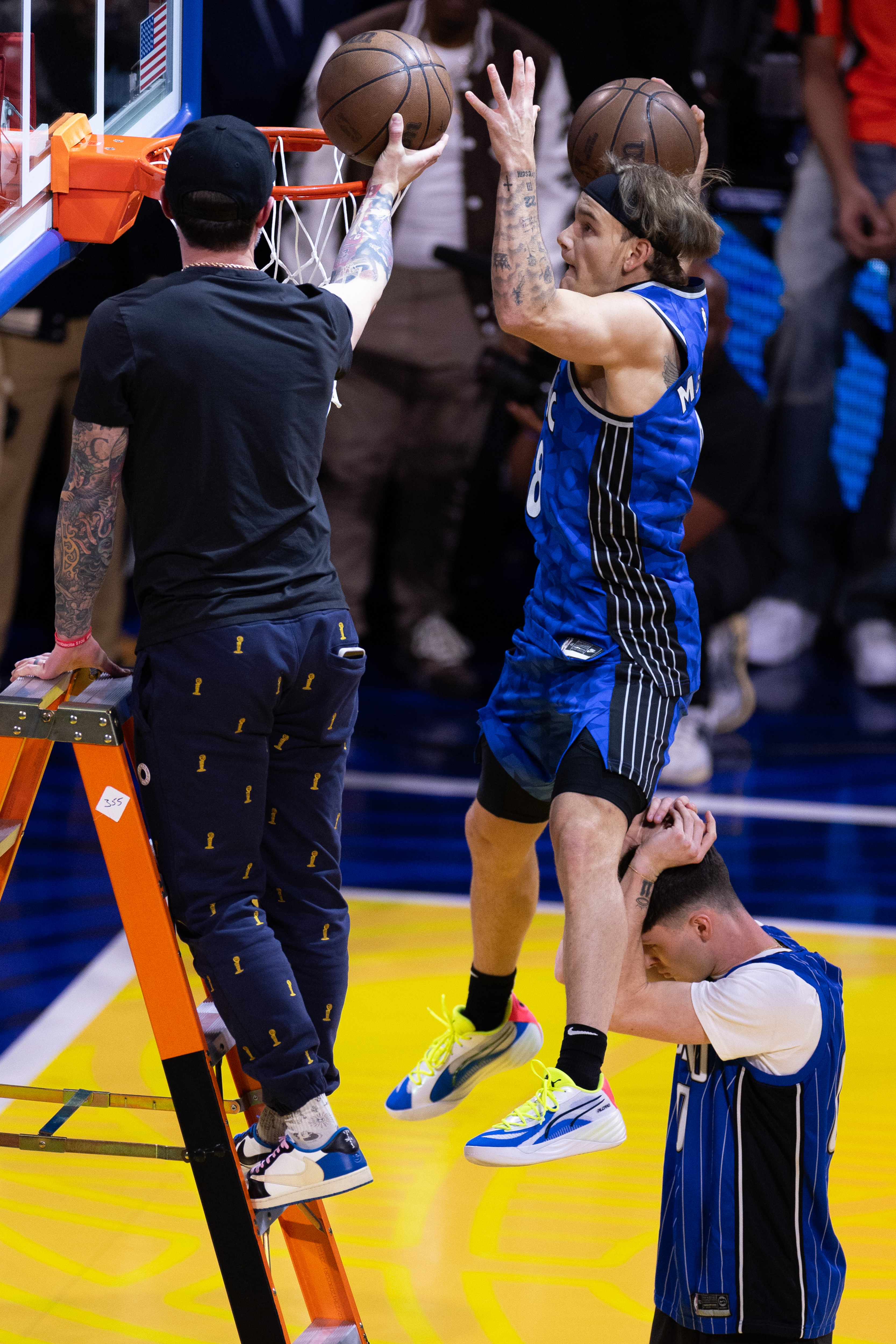 A basketball player in a blue jersey jumps, supported by a teammate holding his legs. He is reaching to dunk a ball, while another person stands on a ladder.