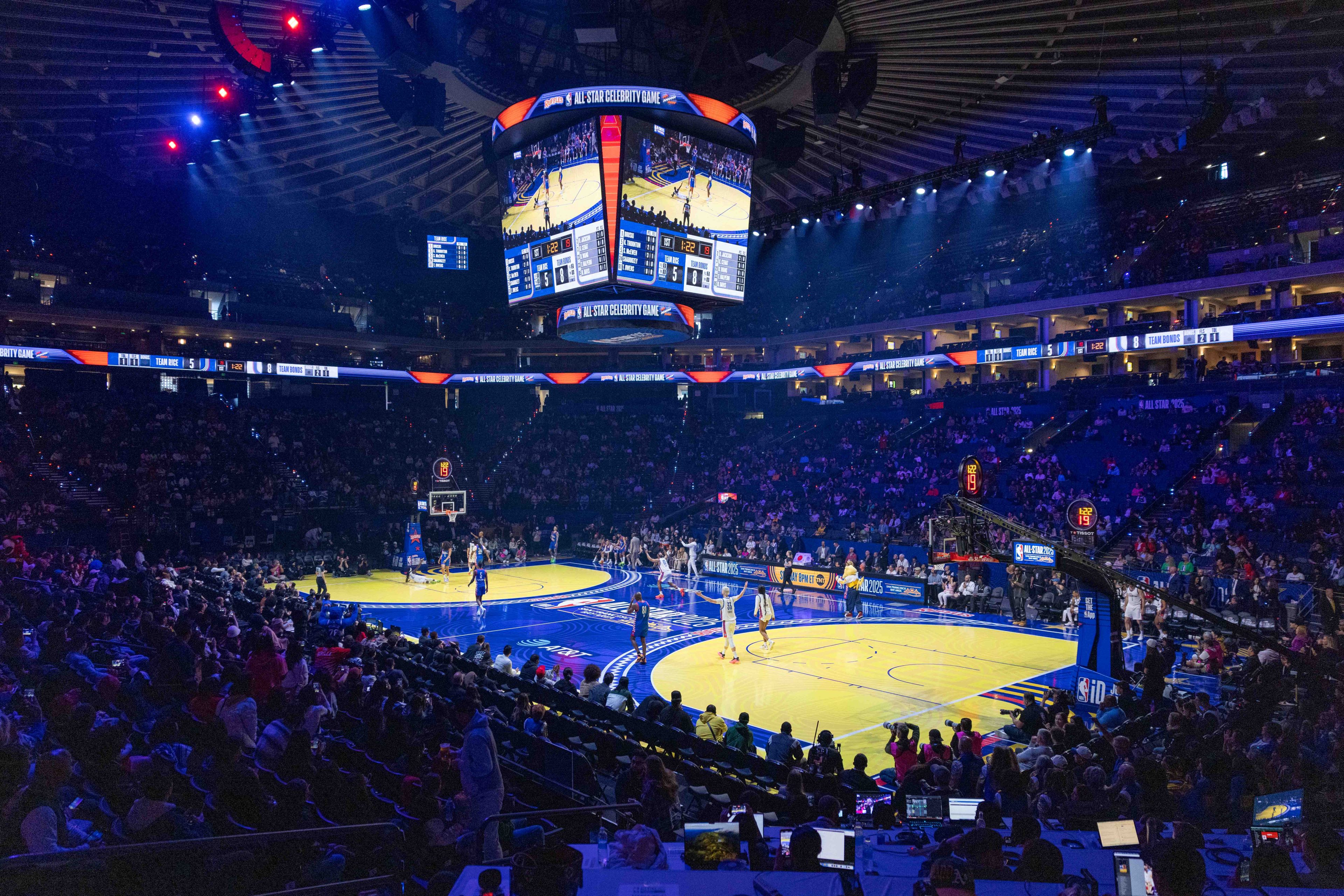 The image shows a large basketball arena during a game, with players on the court and a scoreboard above. The audience is seated around, under dim blue lighting.