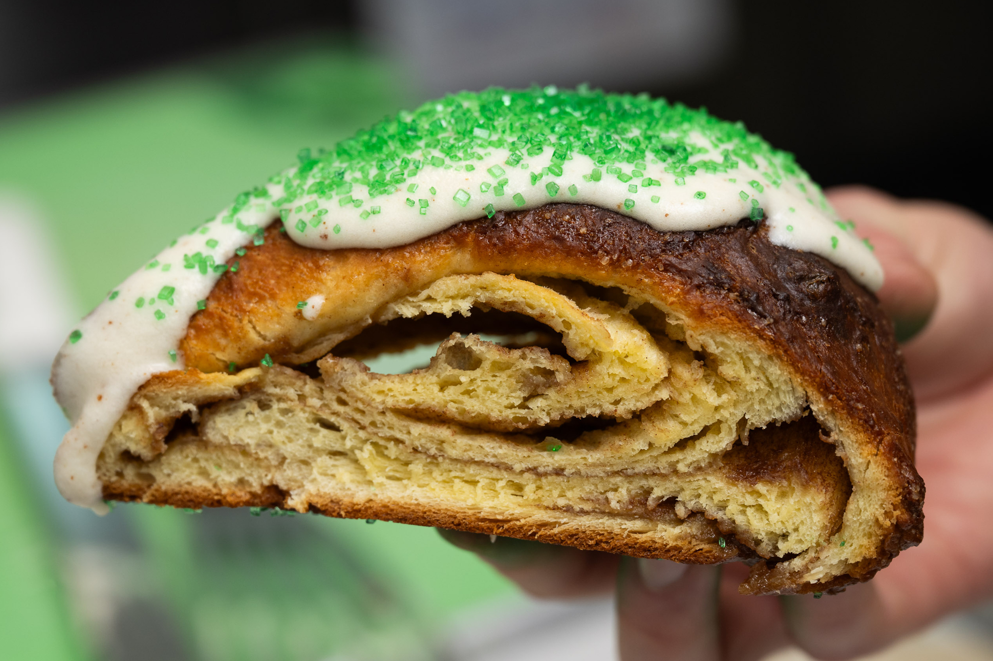 A slice of a pastry decorated with white icing and green sprinkles. The interior shows spiraled layers, hinting at a cinnamon or similar filling.