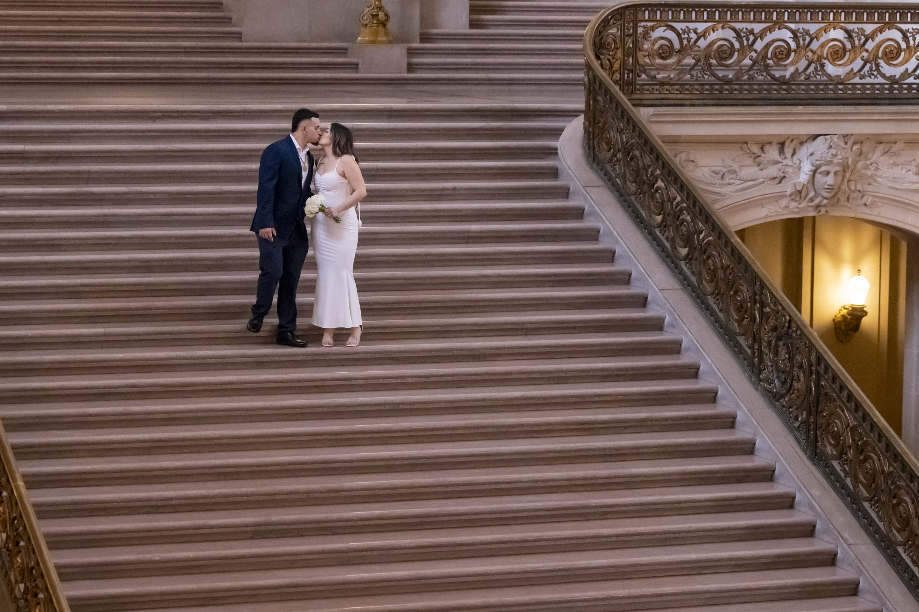 A couple in formal attire shares a kiss on a grand staircase. The setting is elegant and ornate, with intricate railings and decorative walls.