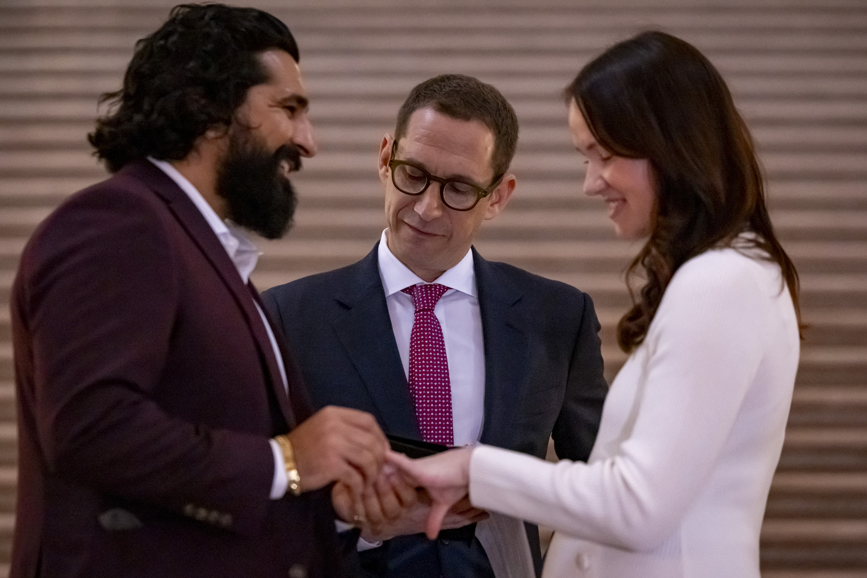 A couple is exchanging rings in a ceremony while a man, possibly an officiant, watches them approvingly. They are smiling and dressed formally.