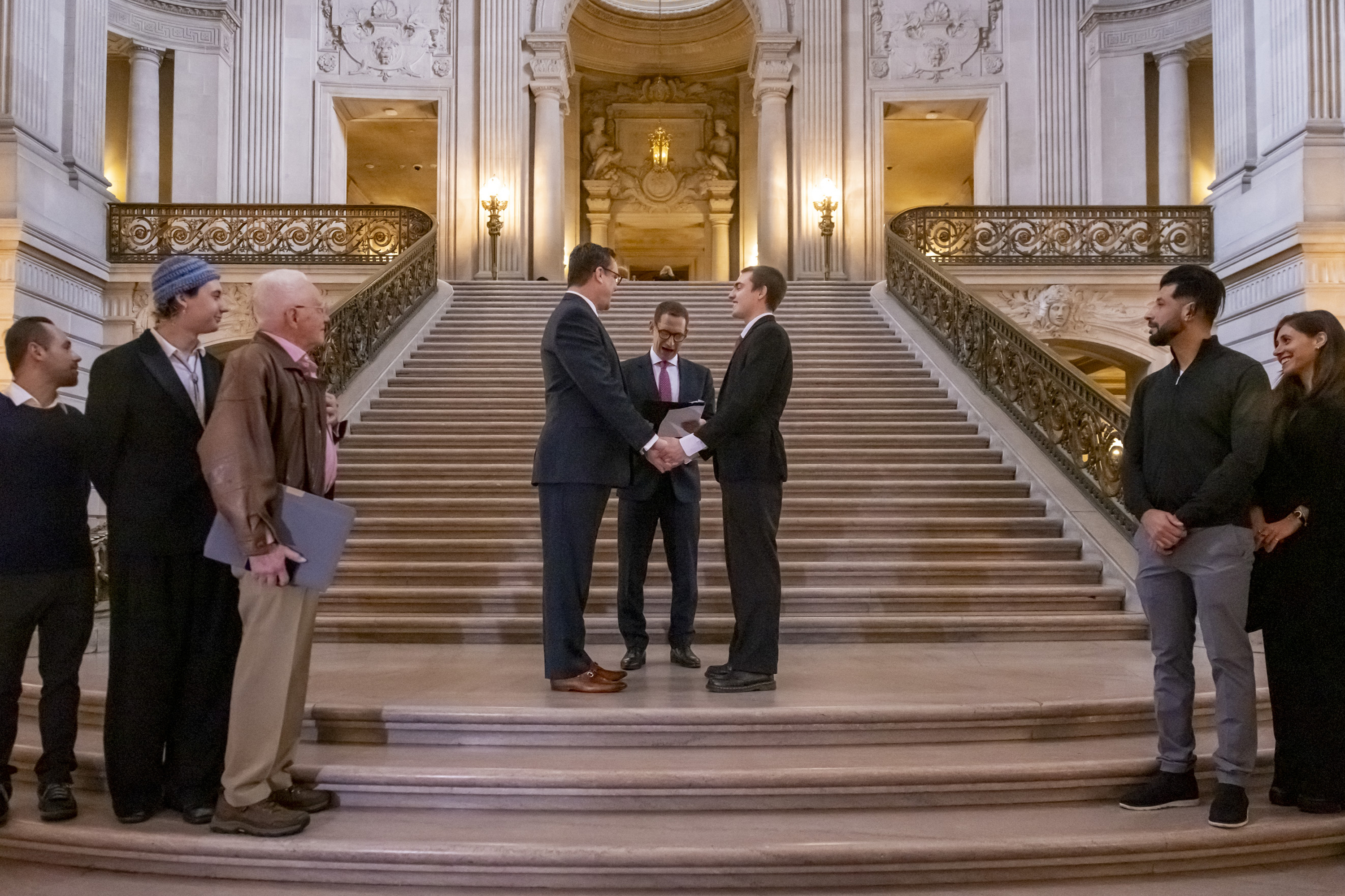 Two people are holding hands on a grand staircase, likely during a ceremony, with a person officiating. Others stand on either side, watching.