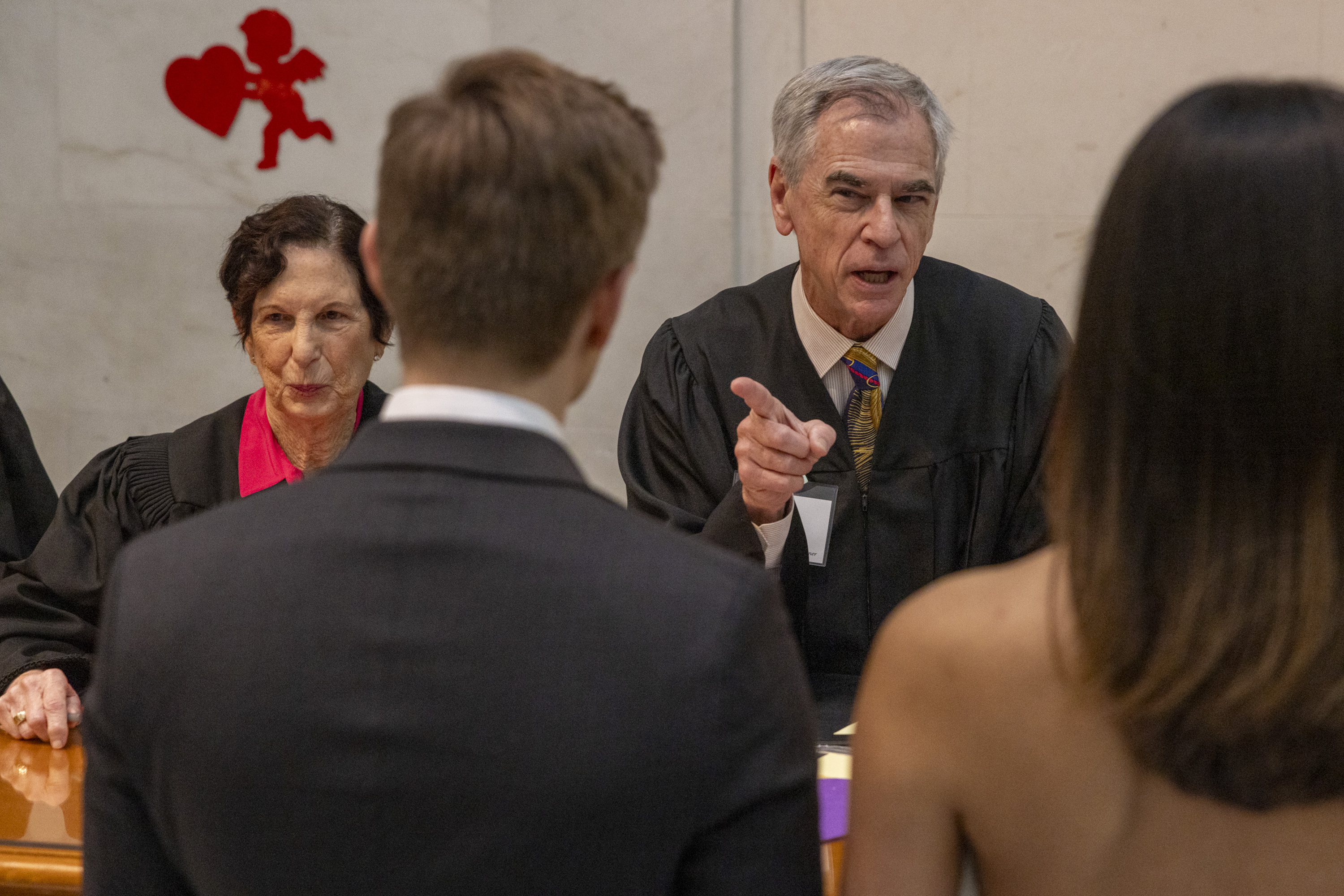 A male judge, wearing a robe and tie, speaks to a couple. A female judge sits next to him. A red cupid with a heart decorates the wall behind them.