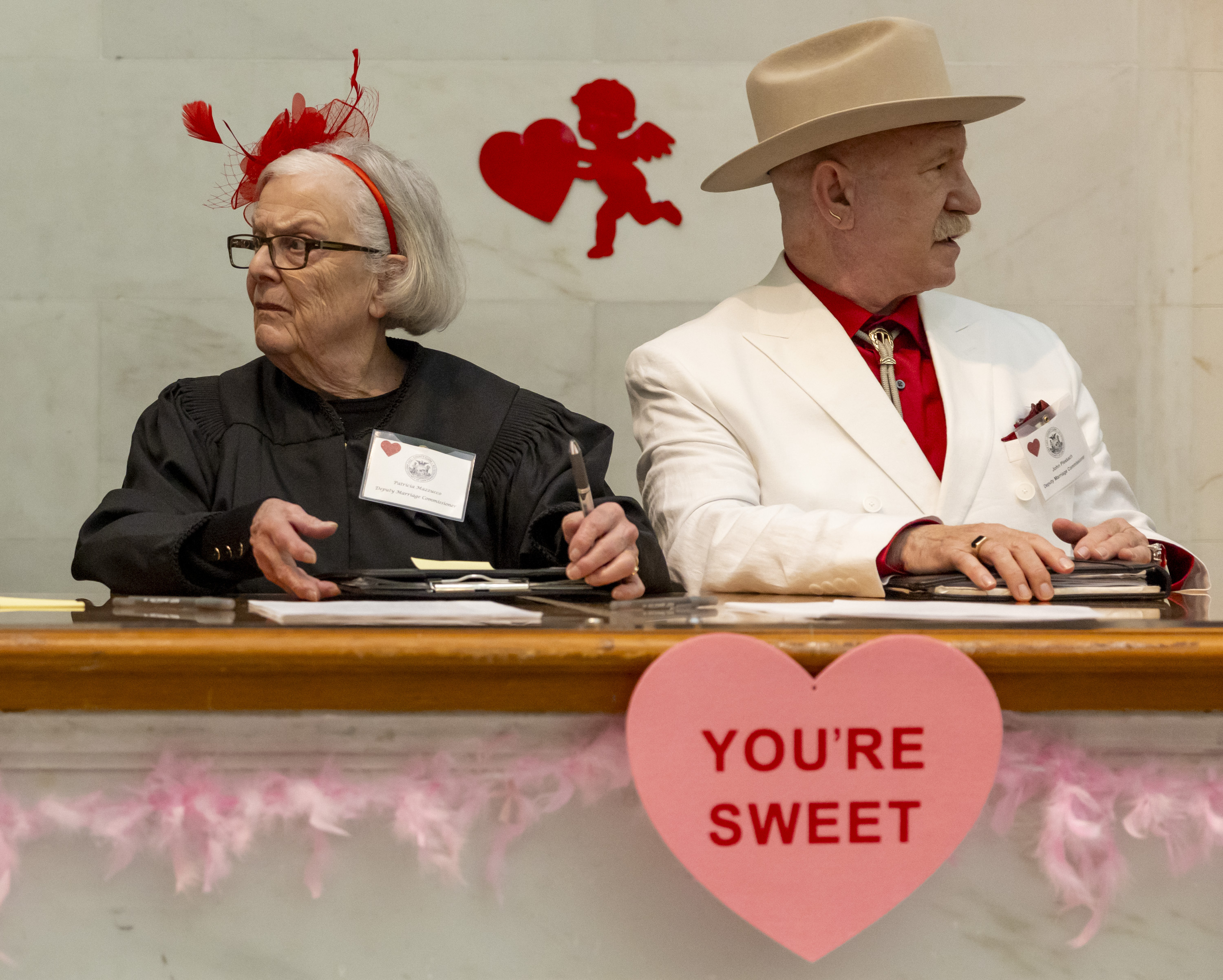 Two people sit at a table, one in a black robe with a red hat, the other in a white suit and cowboy hat. There are heart decorations and a sign saying &quot;YOU'RE SWEET.&quot;