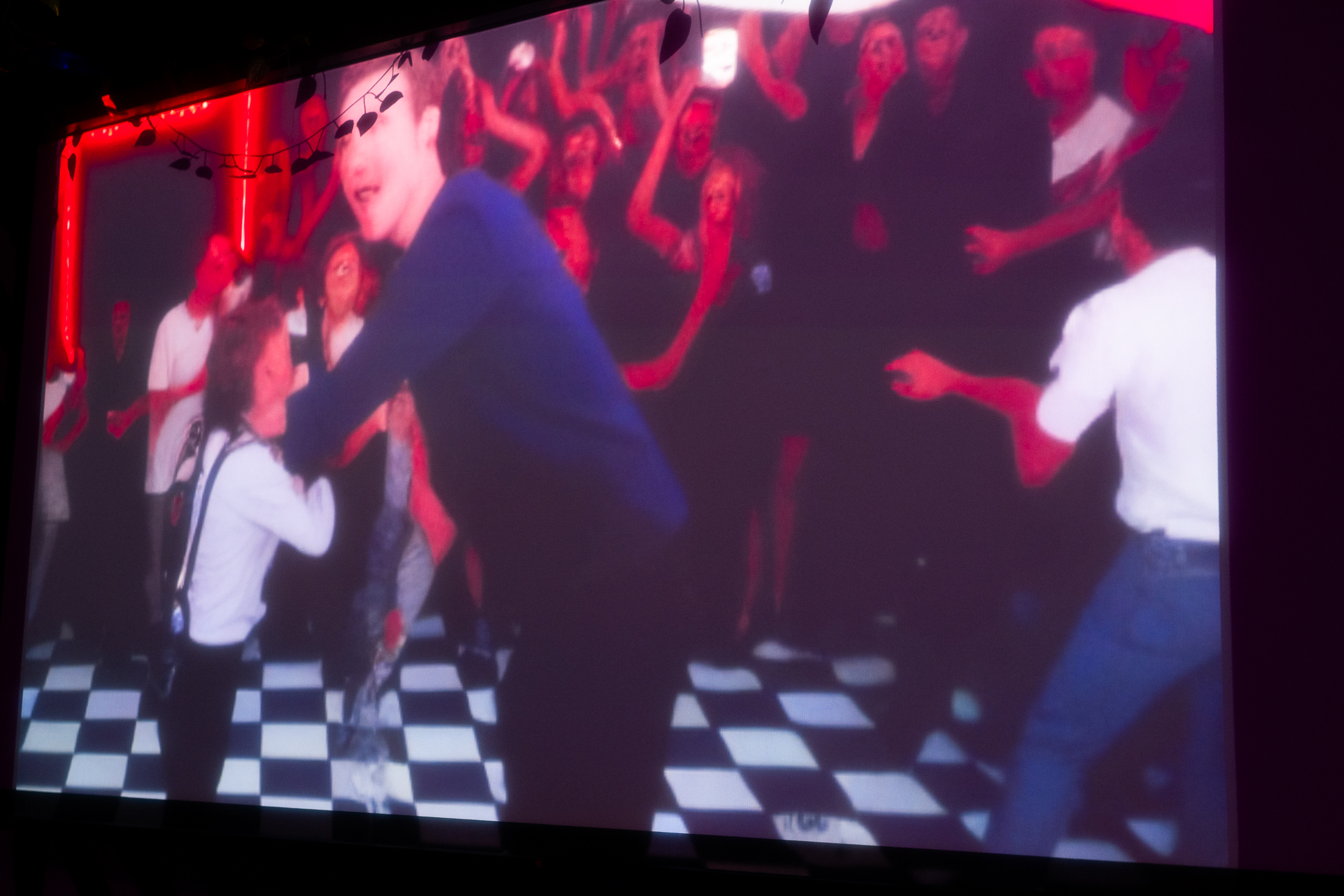 The image shows a lively, blurry scene of people dancing energetically on a checkered floor, surrounded by a vibrant crowd, with red neon lights in the background.