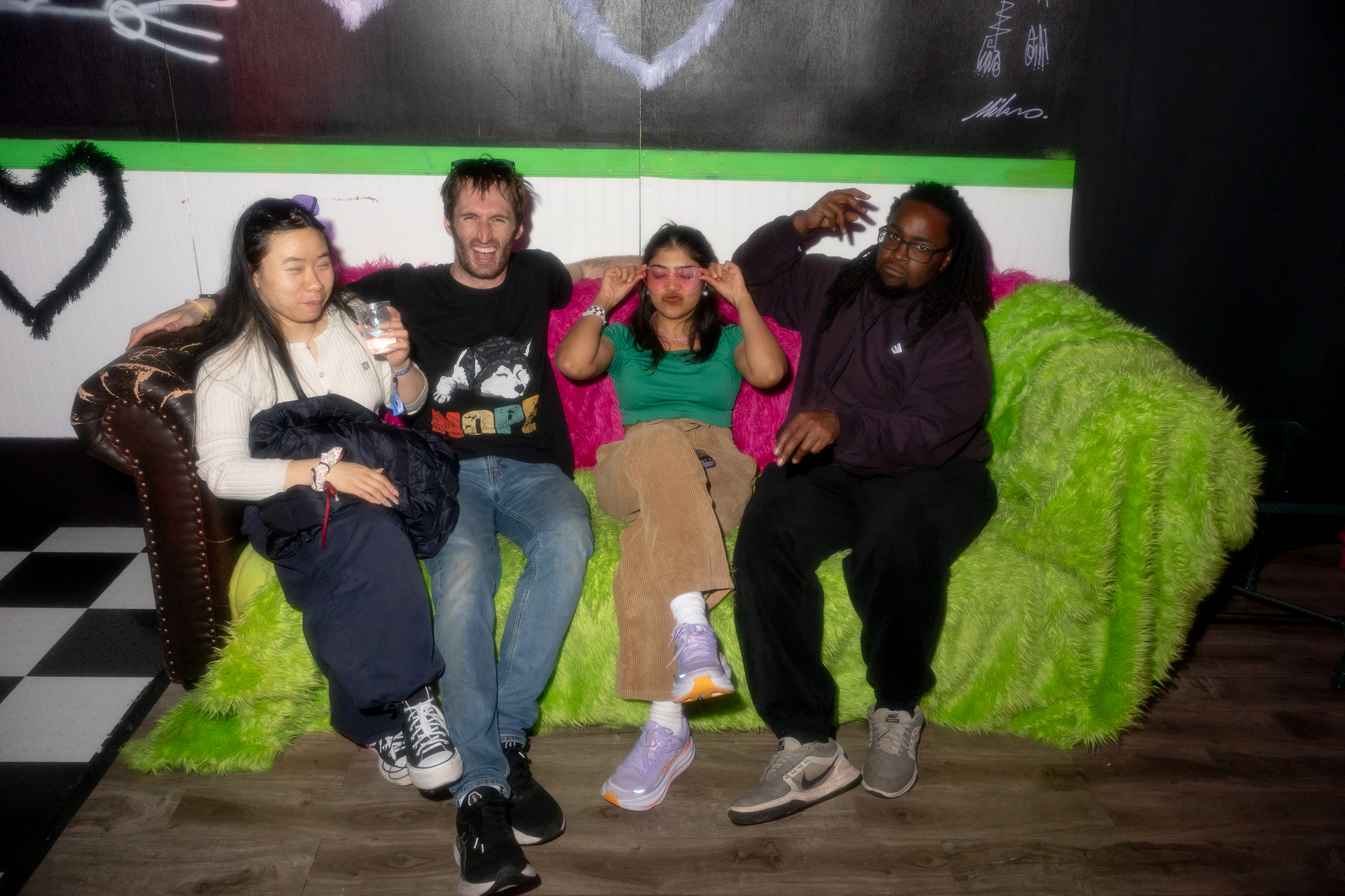 Four people are sitting on a fluffy, bright green couch against a dark wall. One is holding a drink, another is adjusting sunglasses. The mood is casual and friendly.