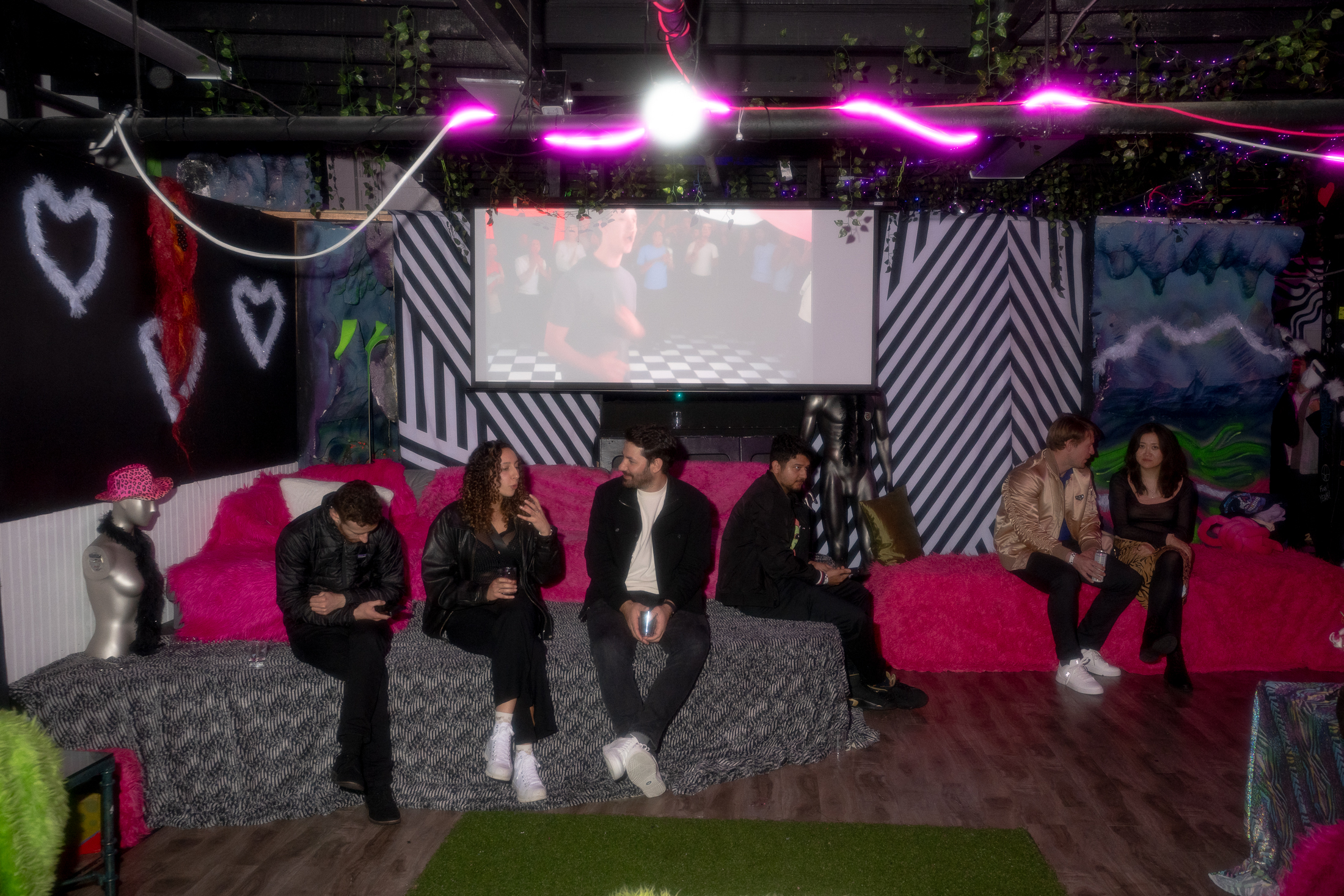 The room features eclectic decor with neon lights and patterns. People sit on colorful couches, and a projector shows a scene. A mannequin wears a pink hat.