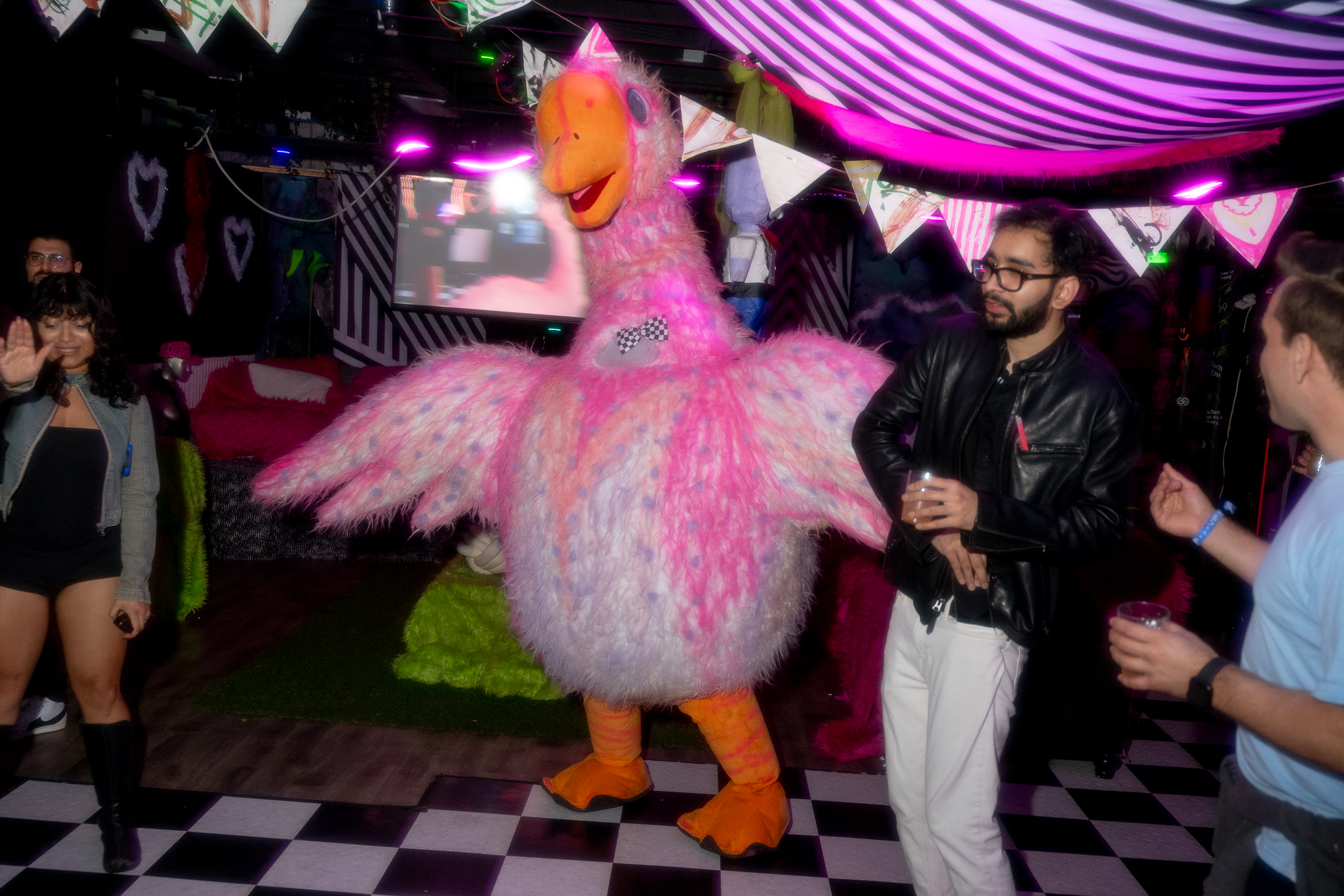 A party scene with a person in a fluffy pink bird costume, people dancing, and colorful decorations under purple lights. The floor is checkered black and white.