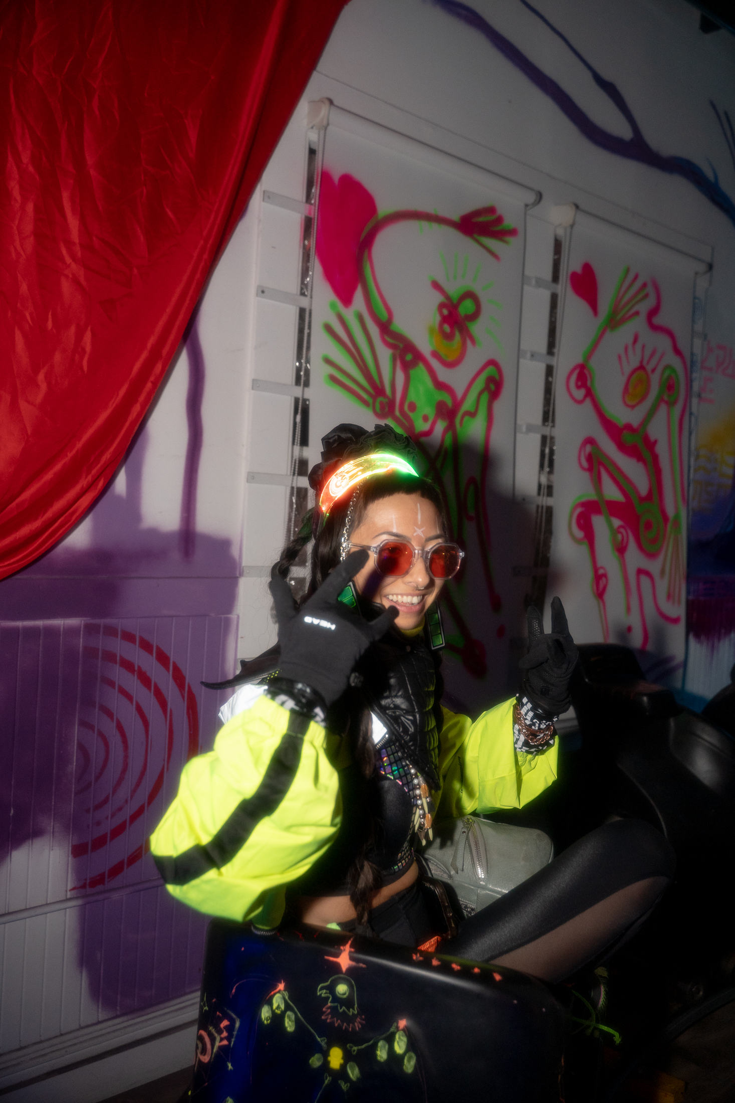 A person in colorful rave attire smiles, wearing LED accessories and glasses, gesturing peace signs. The background has vibrant graffiti and a red curtain.