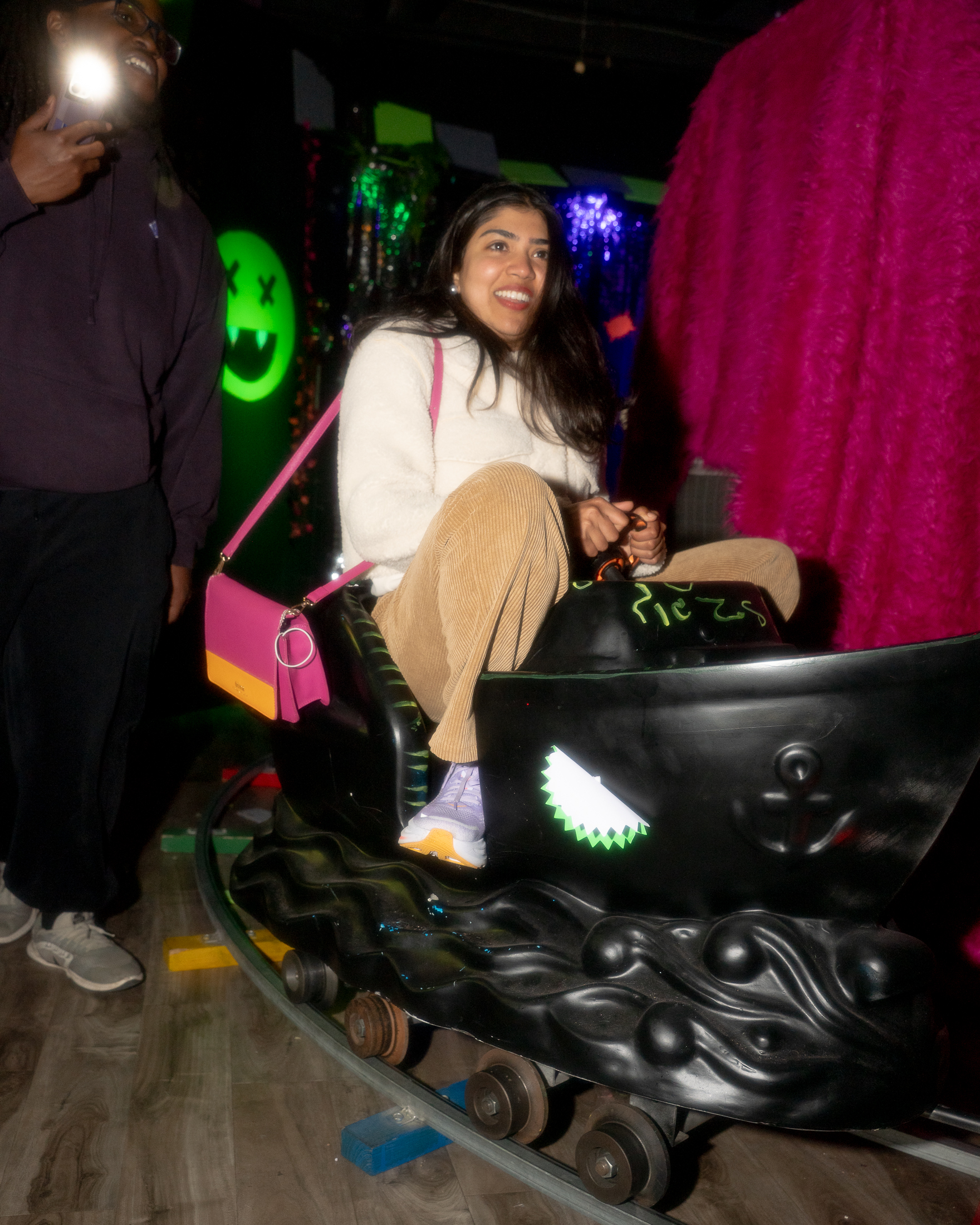 A woman smiles while riding a small, black, boat-shaped amusement ride. Nearby, a person in a hoodie takes a photo. The background is colorful and vibrant.