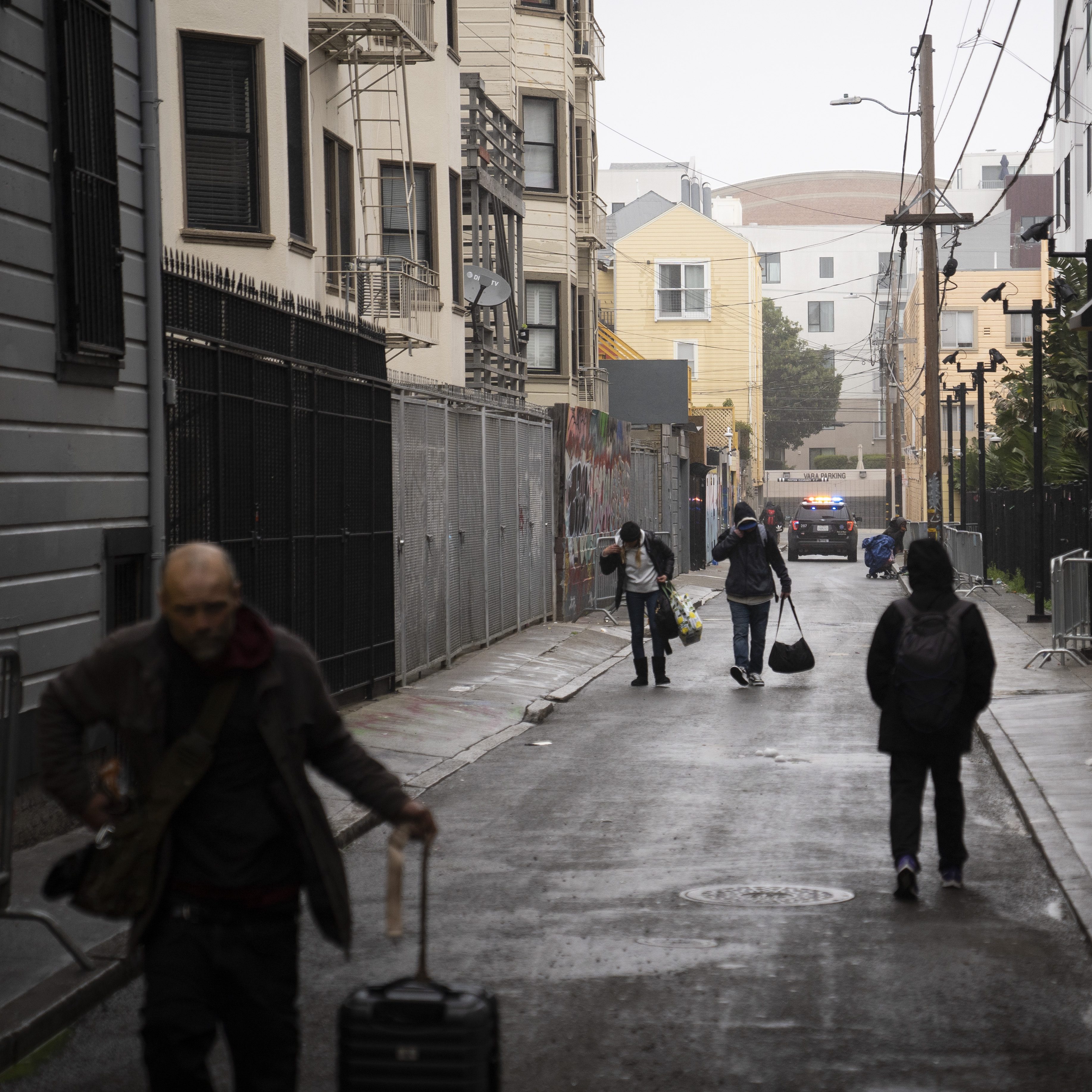 The image shows a narrow alley with people walking, some pulling luggage. Buildings line both sides, and there's a police car in the distance.