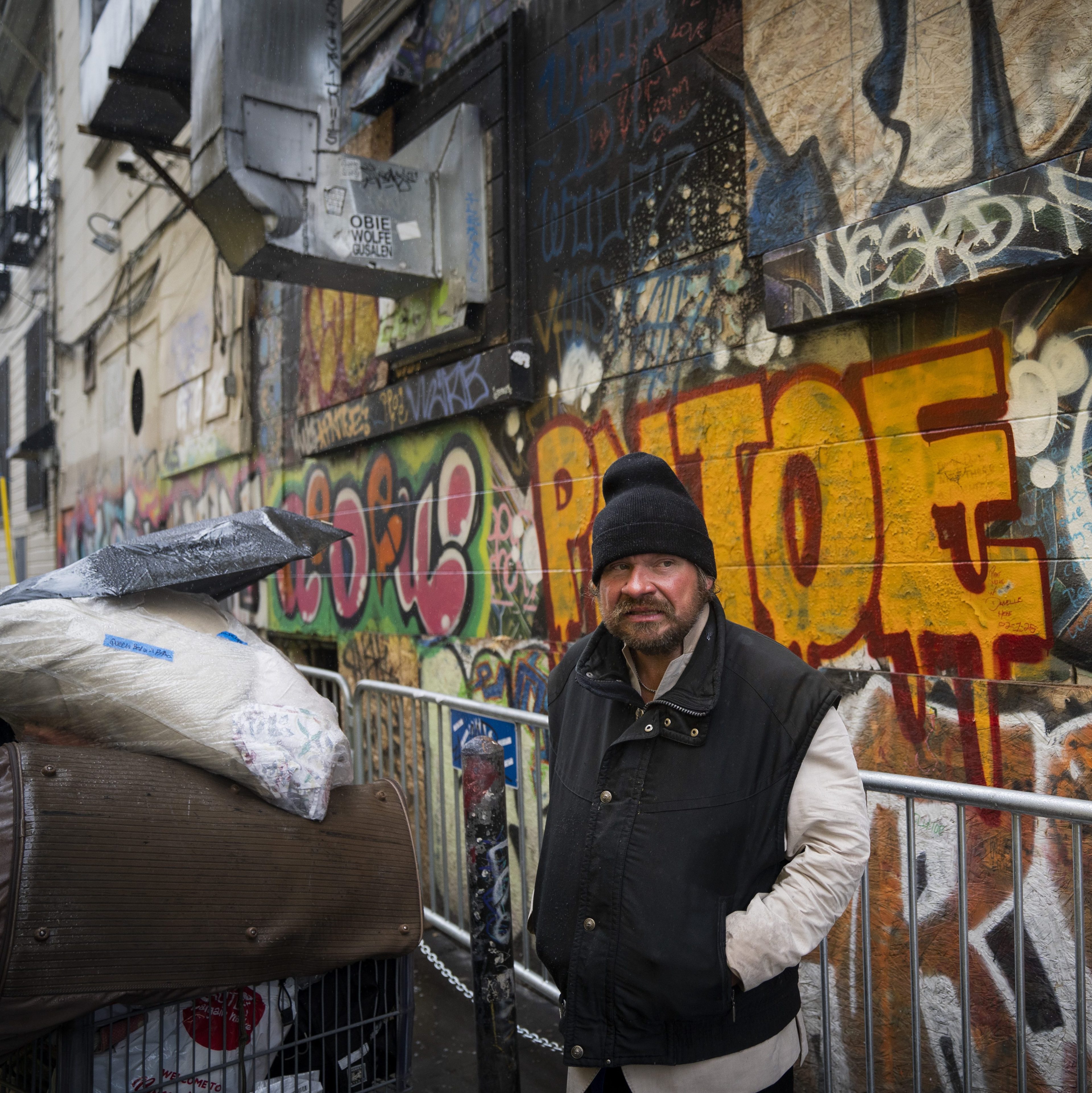 A man wearing a black beanie and jacket stands in front of a graffiti-covered wall, next to a shopping cart covered with a tarp. A &quot;Public Works&quot; sign is visible.