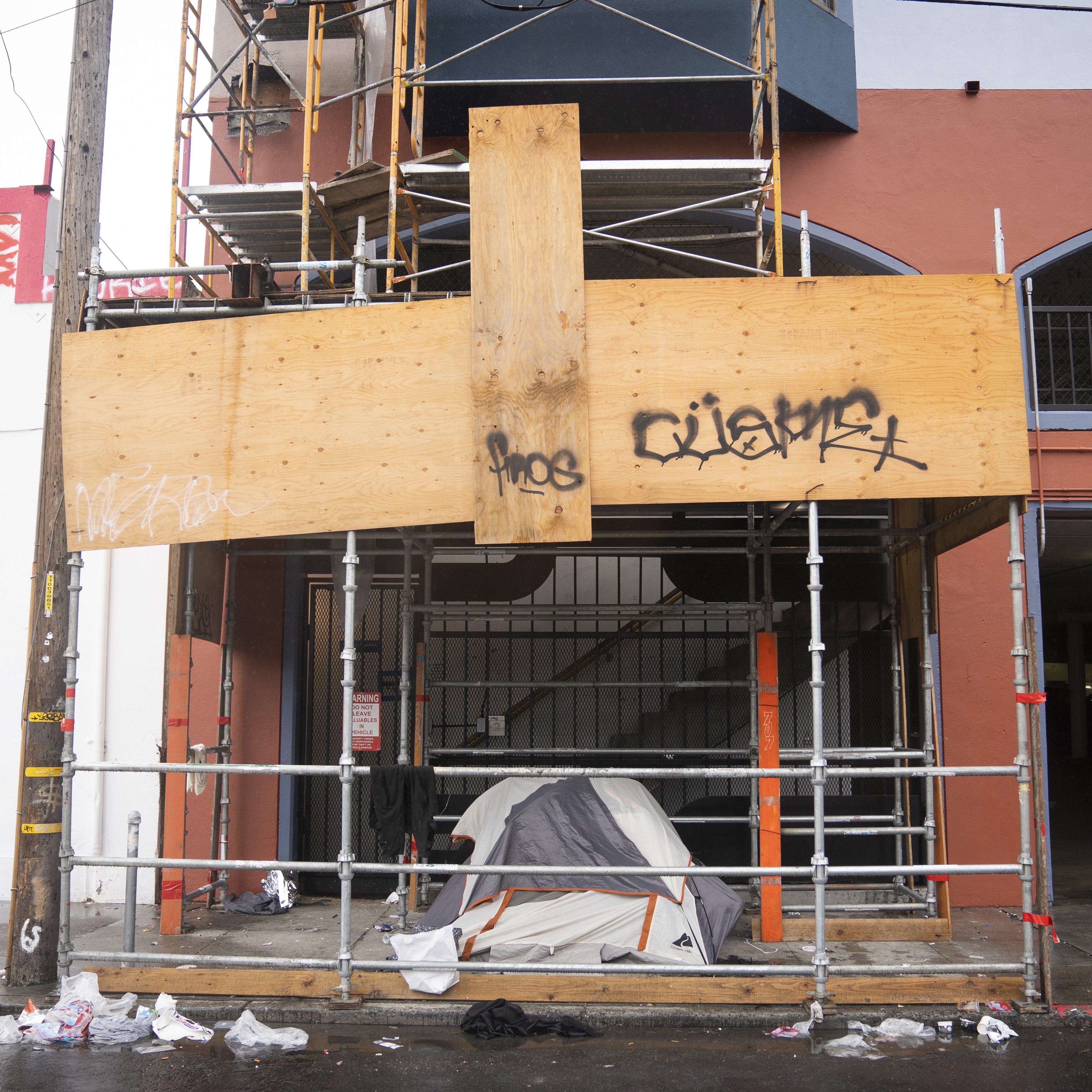 A small tent is set up under scaffolding in front of a building with graffiti. Trash is scattered on the wet sidewalk nearby.