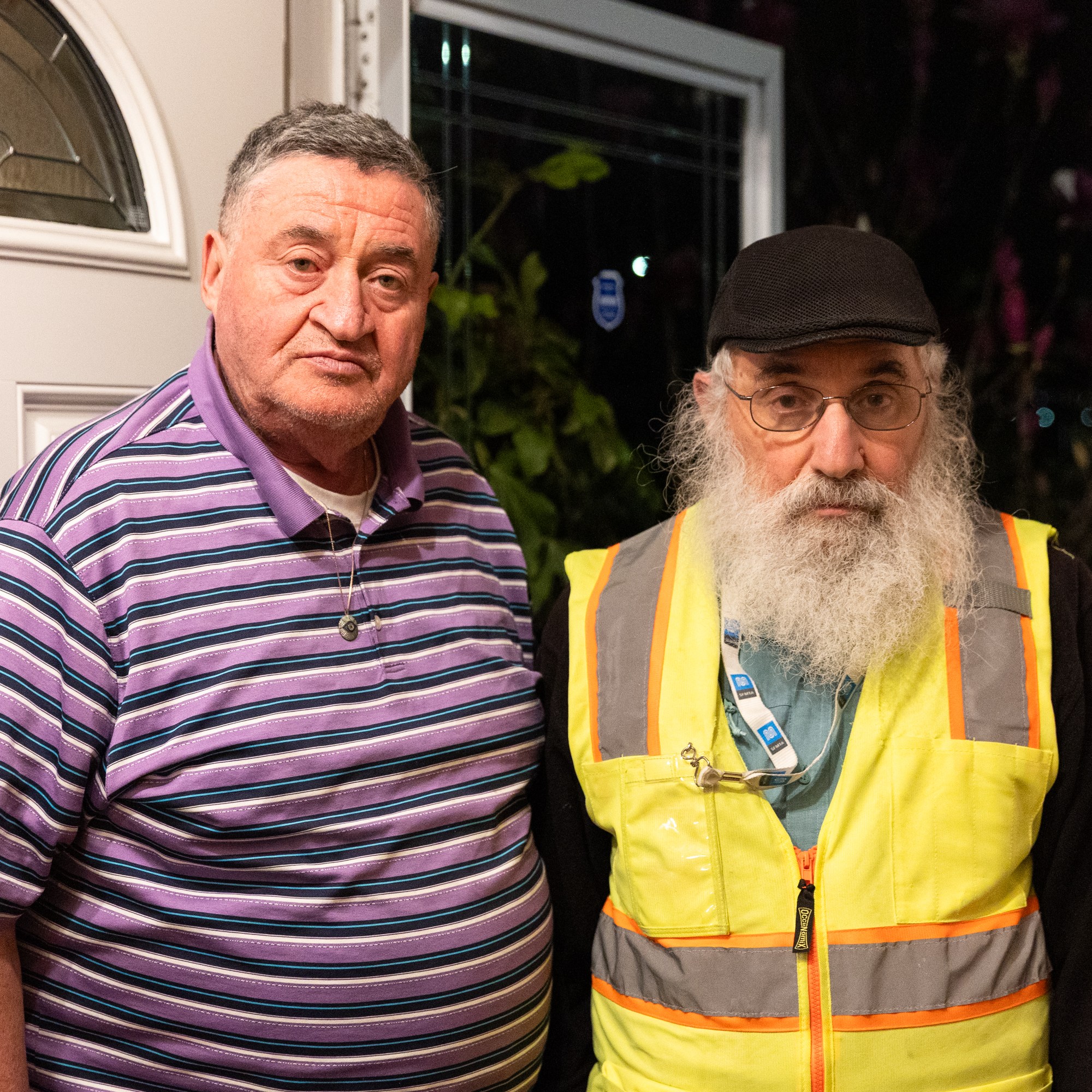 Two older men stand at a doorway. One wears a purple striped polo, and the other dons a black cap with a yellow reflective vest.