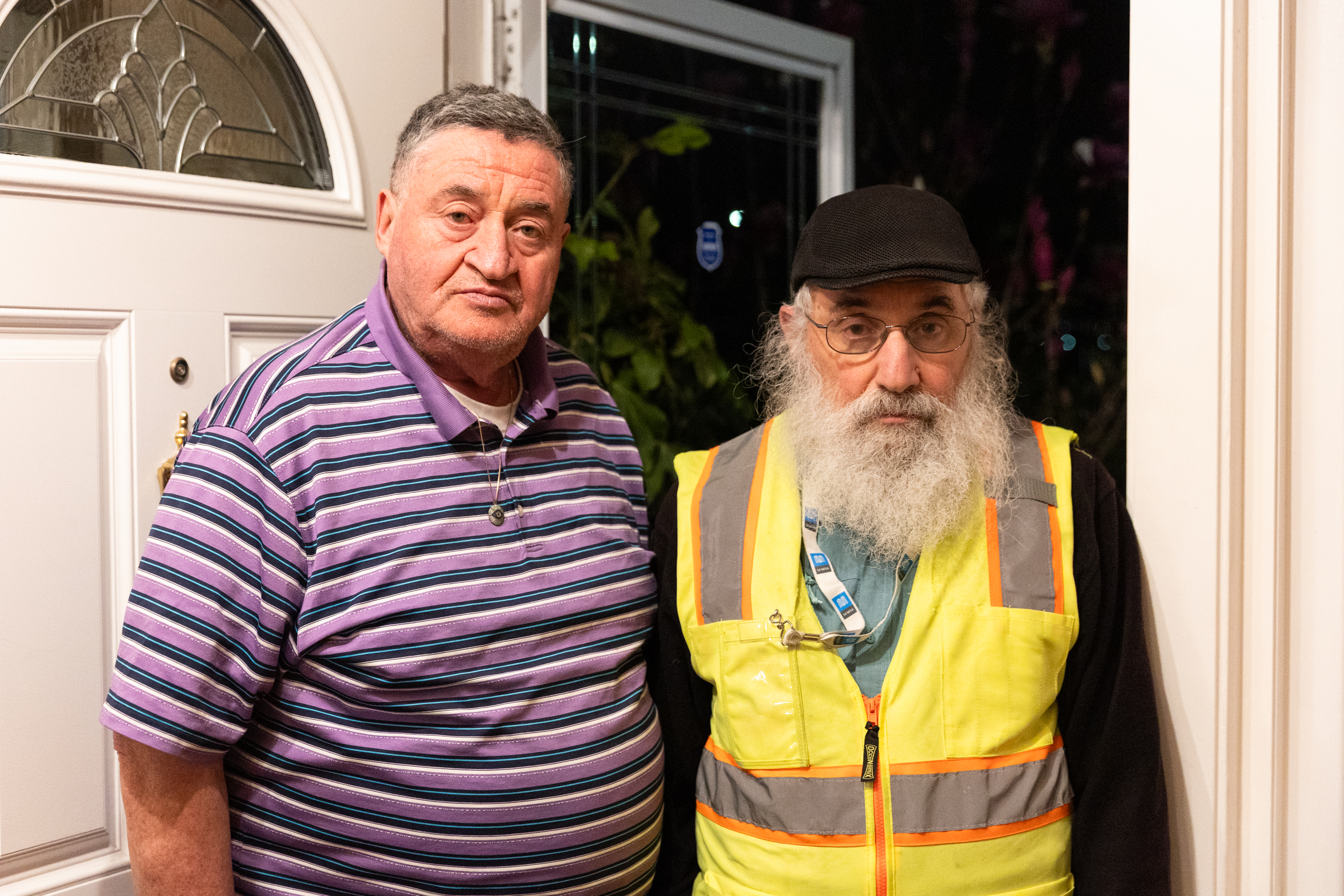 Two older men stand at a doorway. One wears a purple striped polo, and the other dons a black cap with a yellow reflective vest.