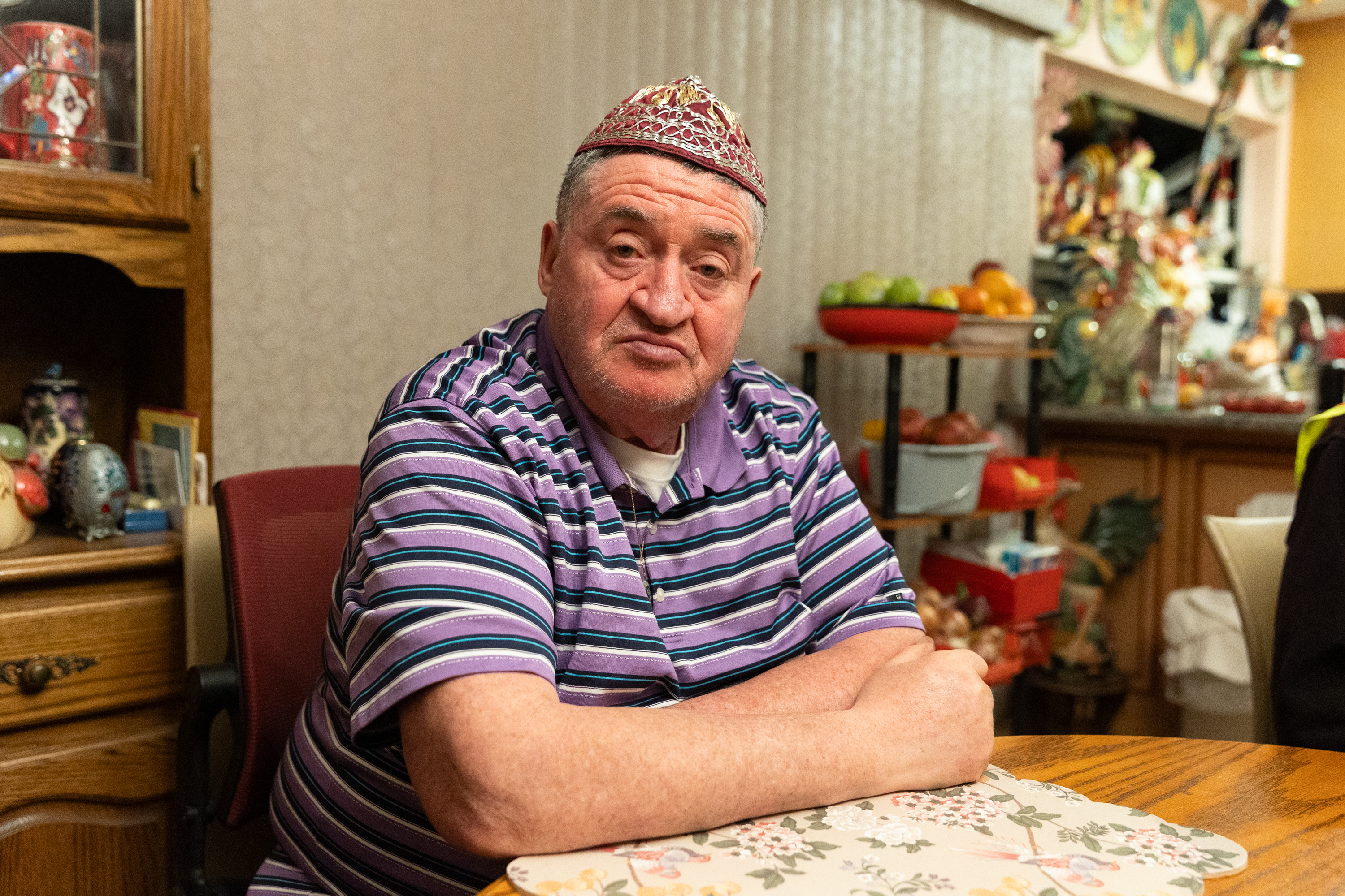 A man wearing a striped purple shirt and a decorative hat sits at a wooden table in a cozy room filled with various decor and fruit.