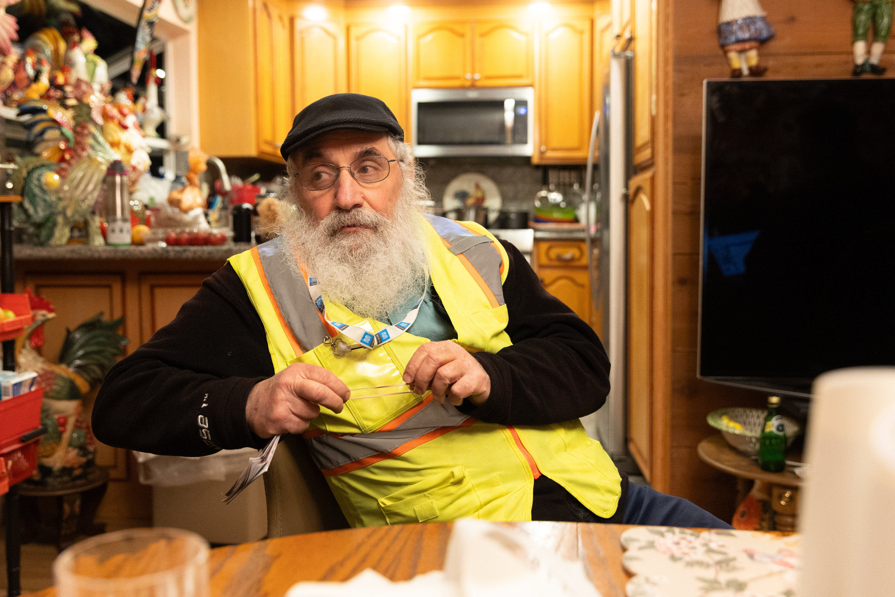 An older man with a beard, wearing glasses, a cap, and a high-visibility vest, sits at a table in a kitchen decorated with rooster figurines.