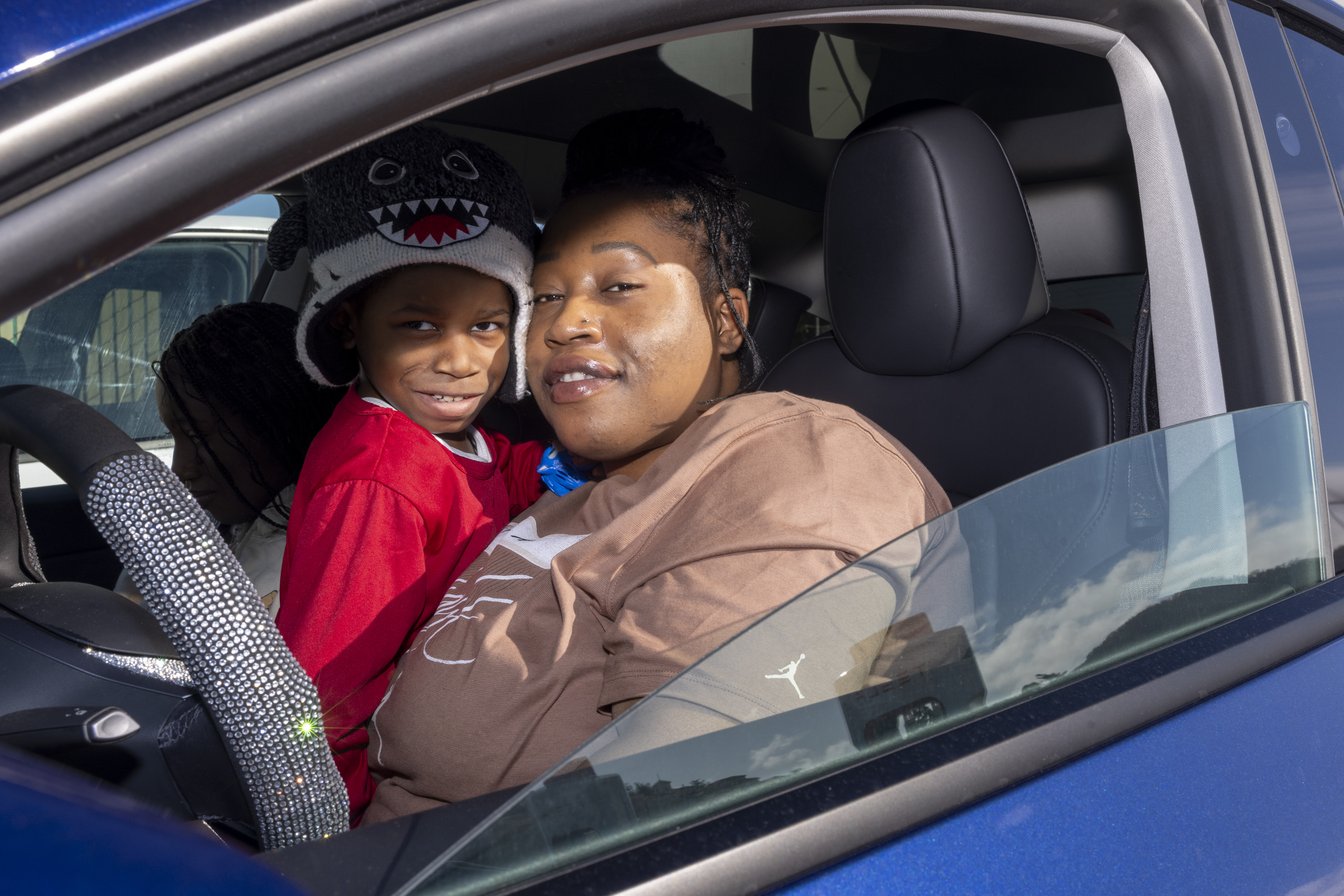 A person is sitting in the driver's seat of a car with a child on their lap. The child is wearing a hat with a shark face. The car's window is open.