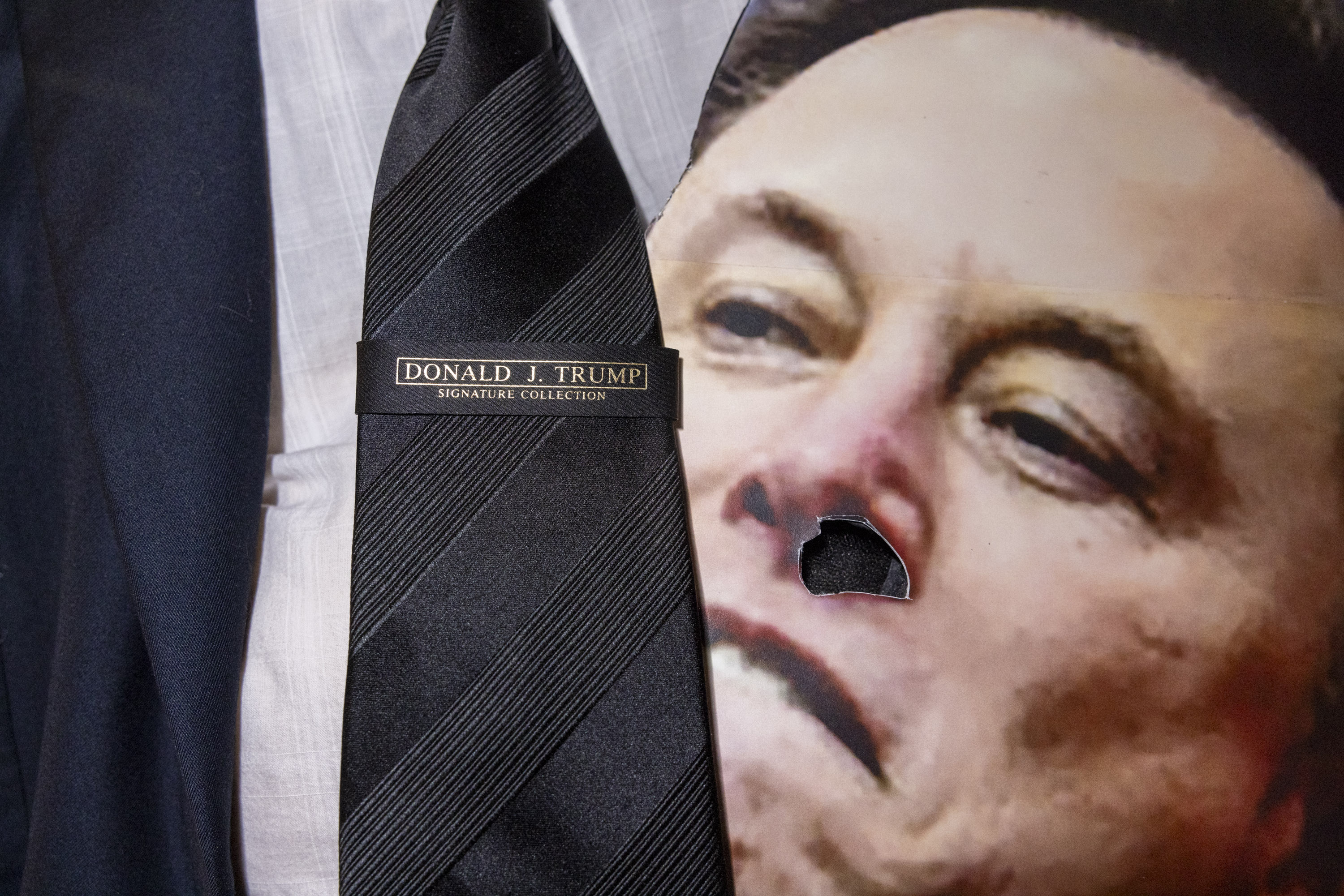 A black tie labeled &quot;Donald J. Trump Signature Collection&quot; is framed against a white shirt and dark suit, with a close-up face image partially shown beside it.