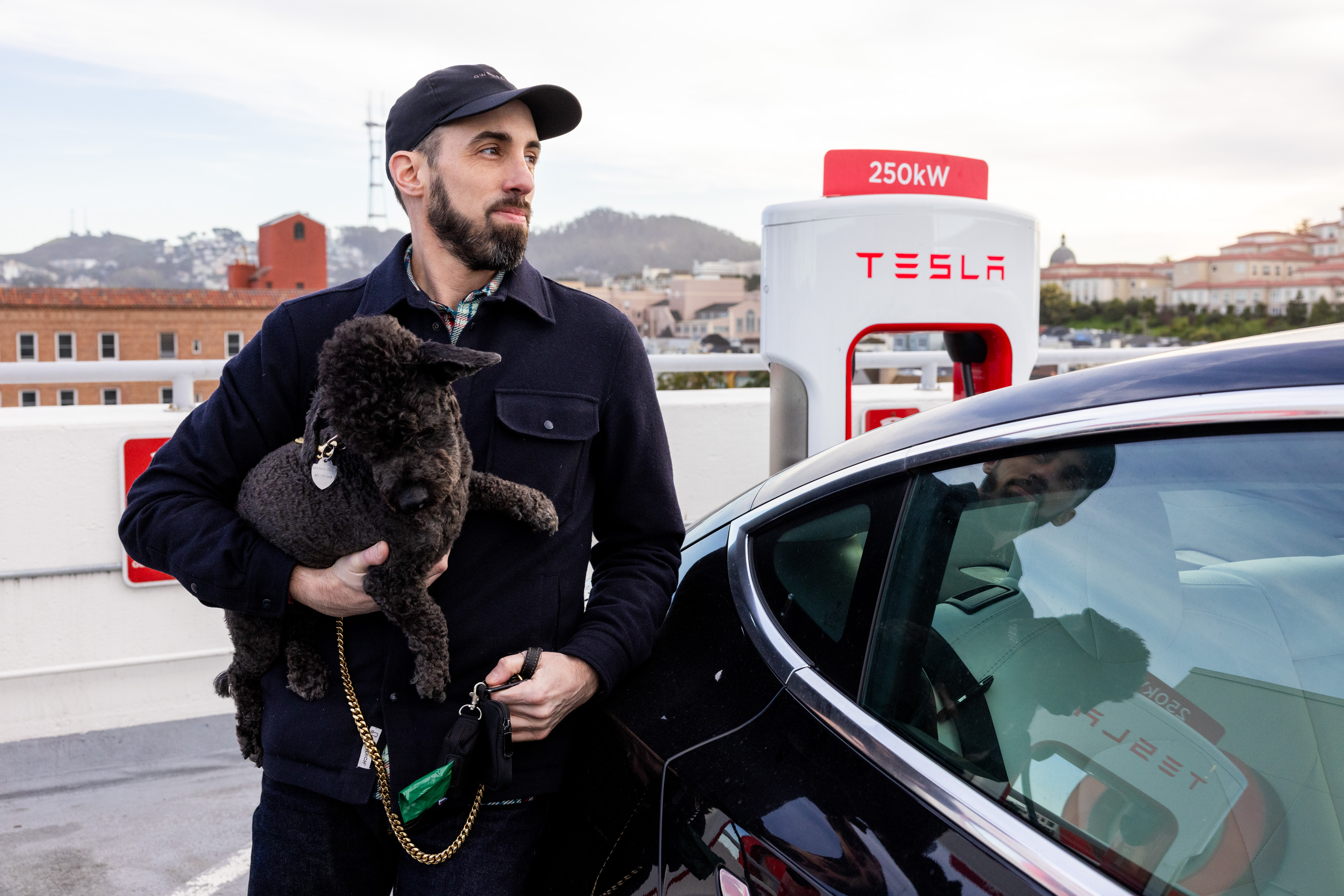 A man holds a black dog beside a Tesla charger. He wears a cap and dark jacket, standing next to a black Tesla. A cityscape is in the background.