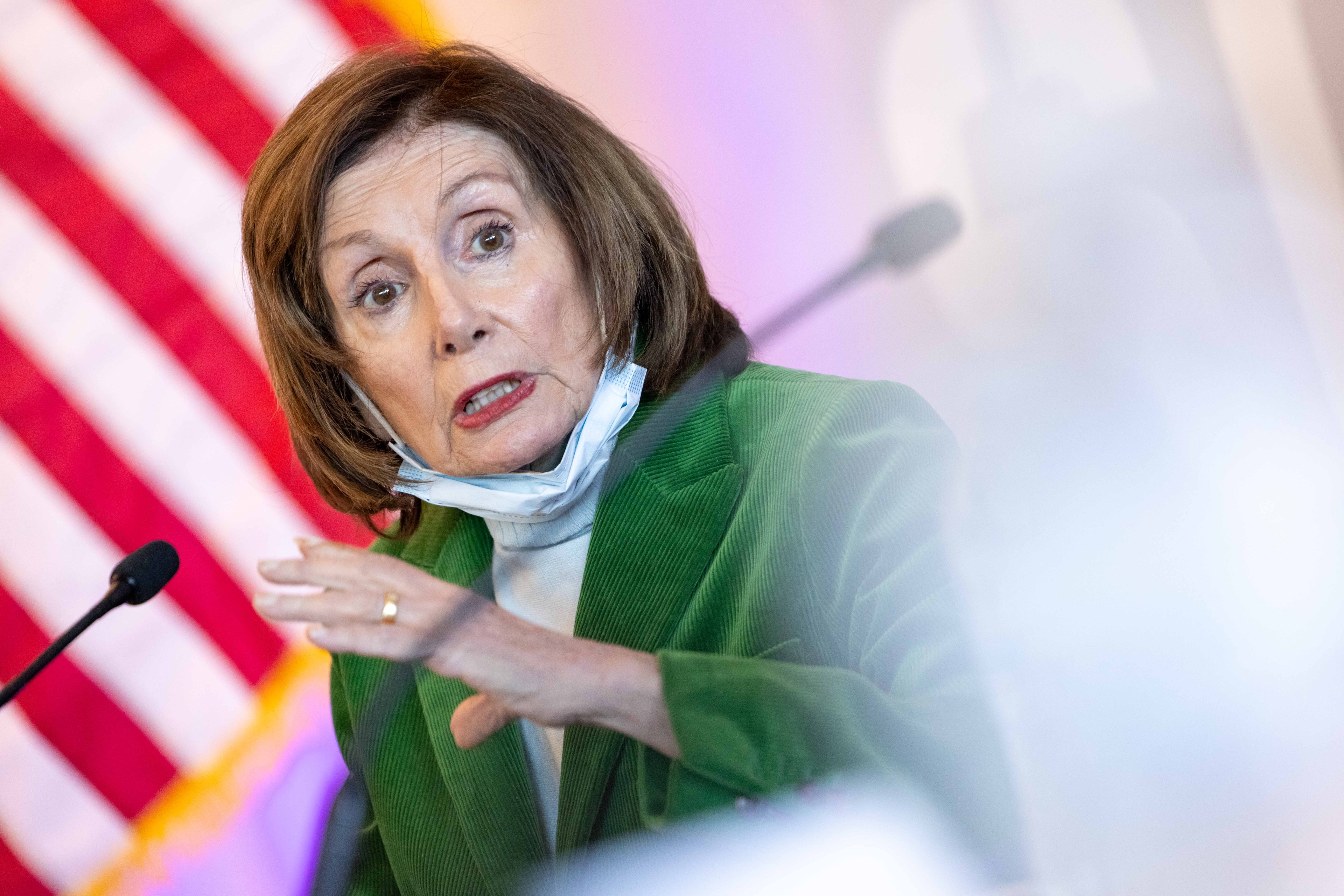 A woman in a green jacket speaks passionately with a microphone nearby, against a blurred background that includes the U.S. flag. She has a mask around her neck.