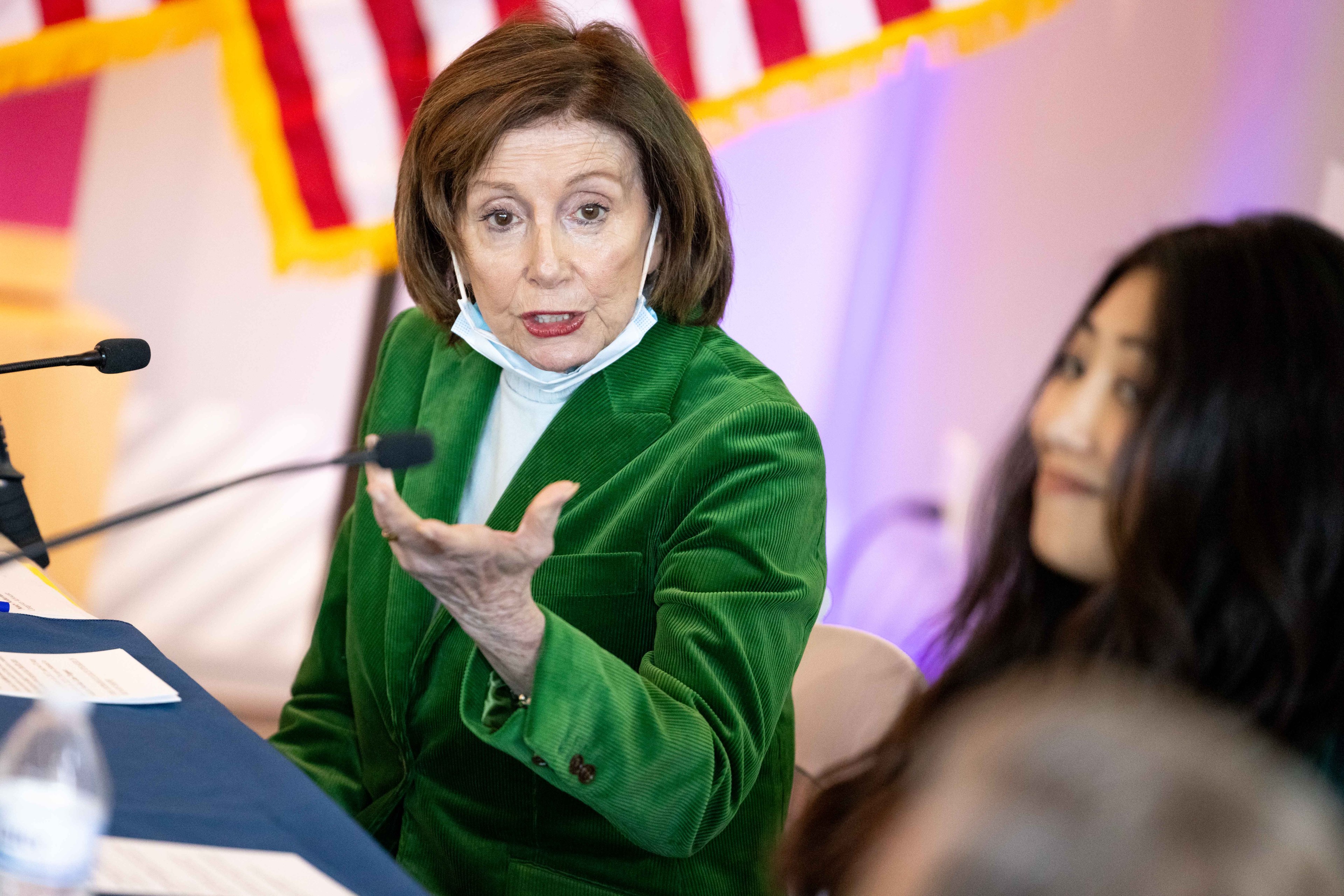 A woman in a green jacket speaks animatedly at a table with microphones, while another person watches. An American flag is partially visible in the background.