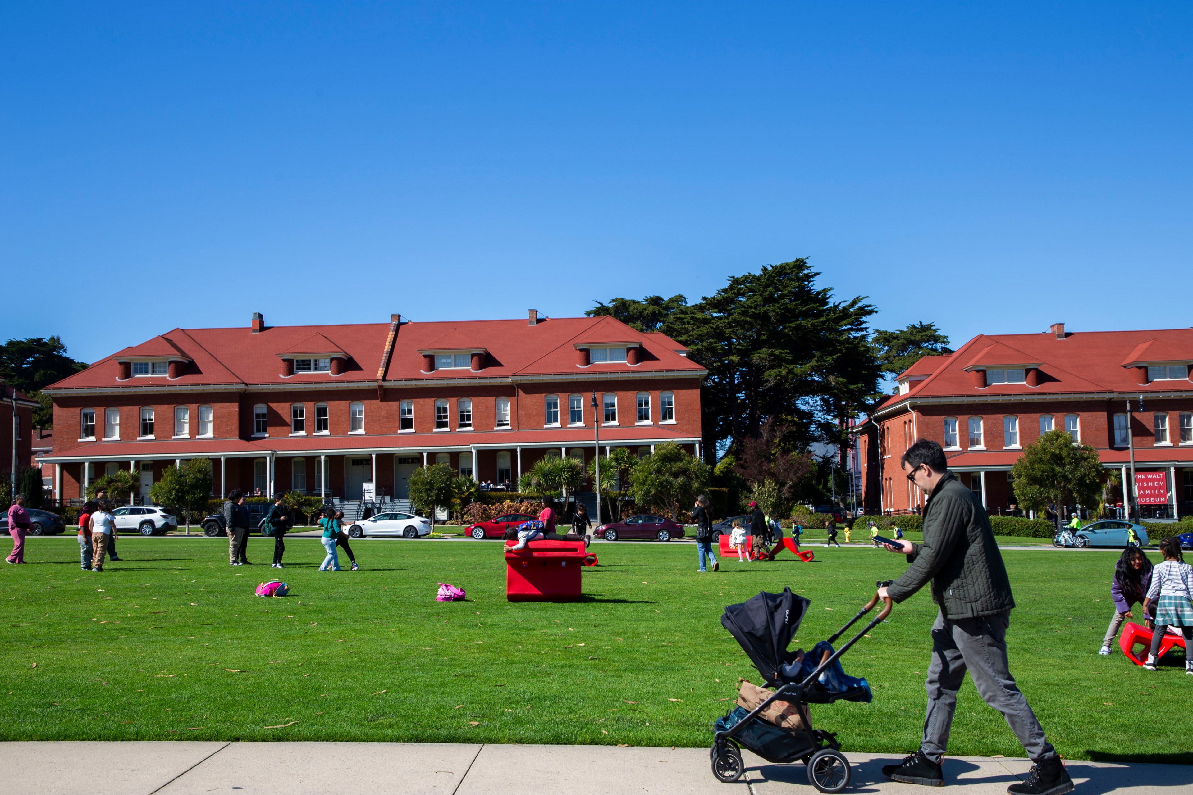 A man pushes a stroller across a grassy field, where people are gathered and children play. Red brick buildings with red roofs line the background under a clear blue sky.