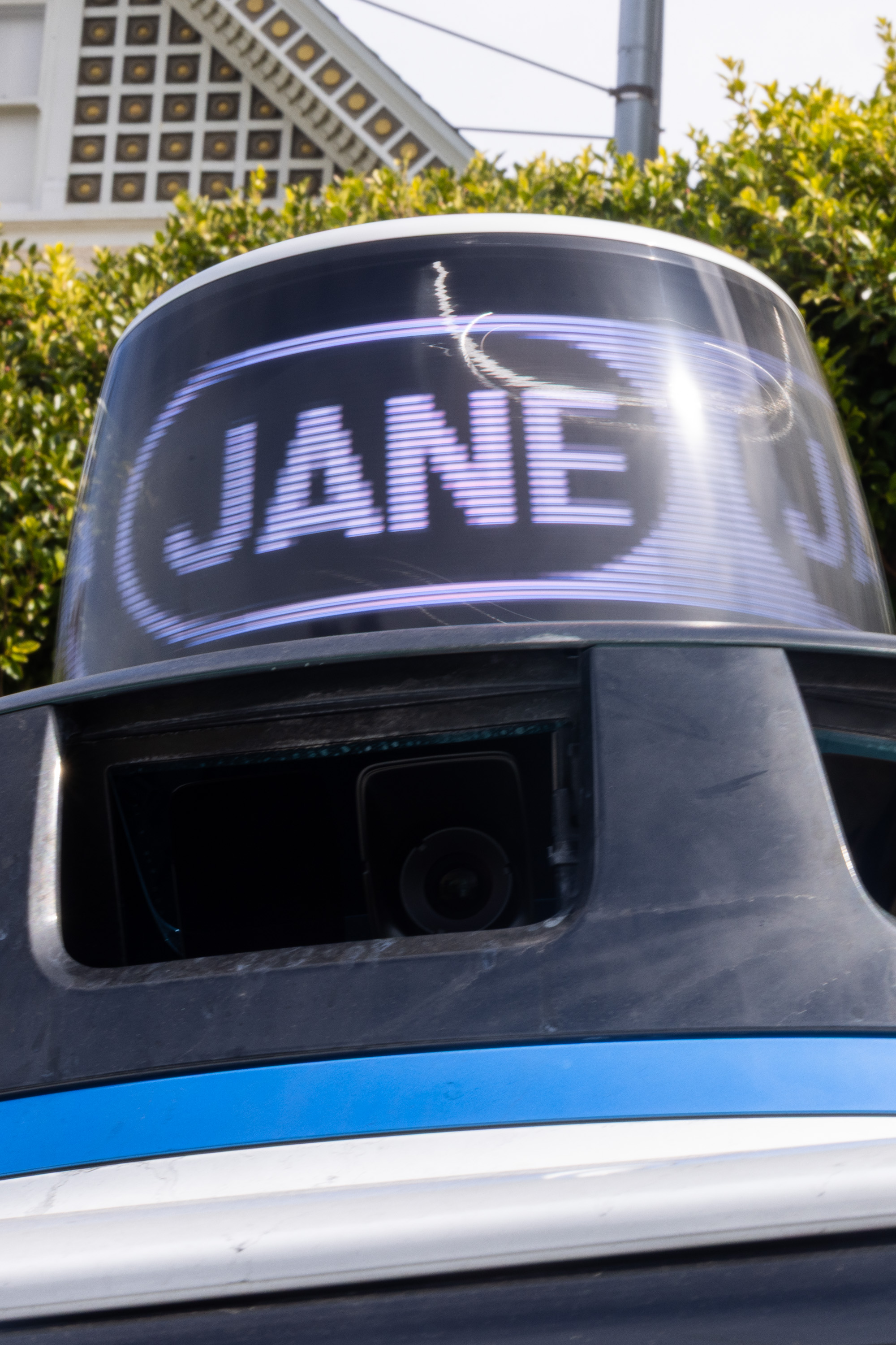 A robotic device is topped with a curved screen displaying the name &quot;JANE.&quot; Behind it, there are bushes and a patterned building facade.