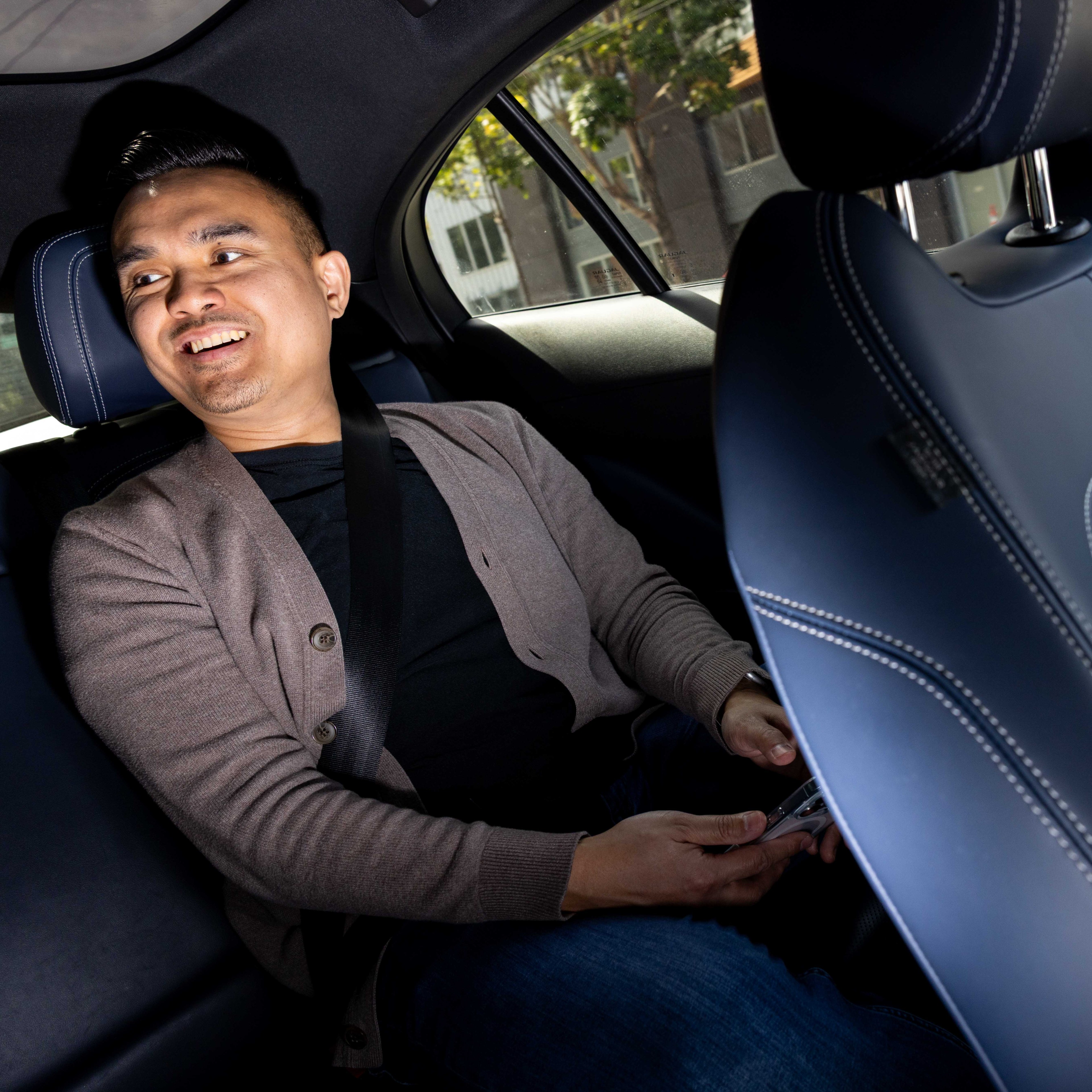 A person wearing a gray cardigan is smiling while seated in the backseat of a car, holding a phone. The car interior is dark with blue accents.
